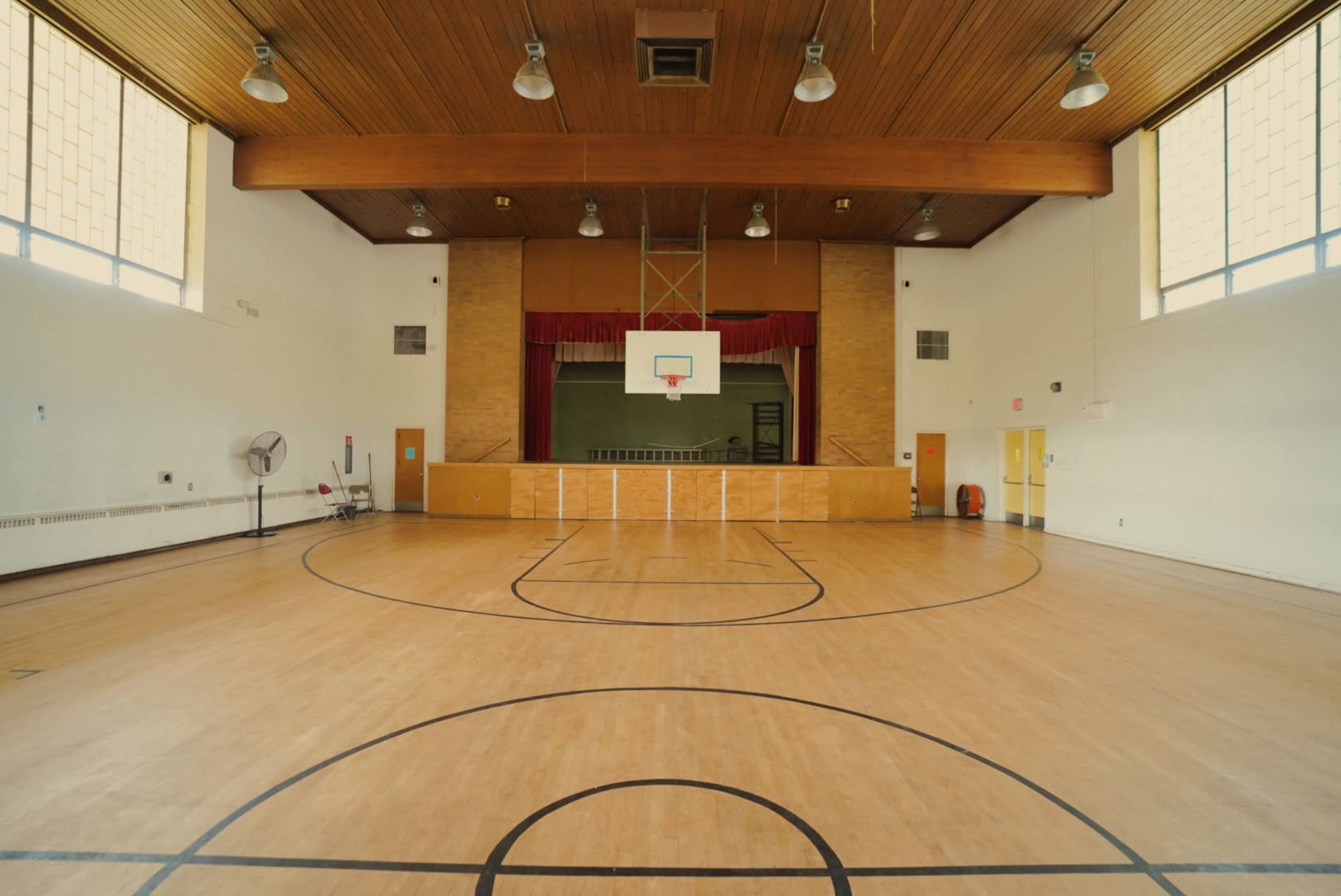 An empty gymnasium with a basketball hoop, a wooden floor, and a stage at the back.