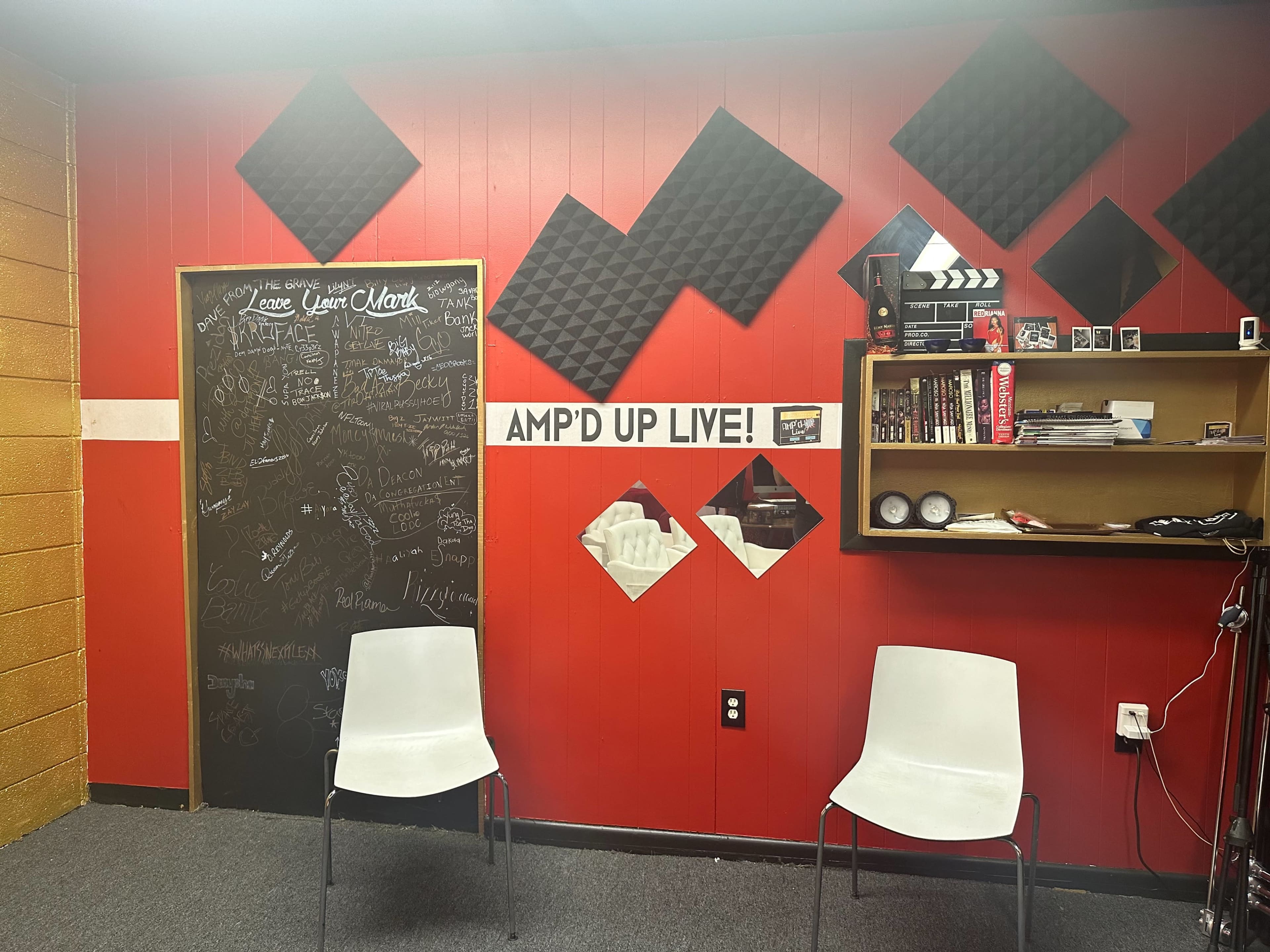 A room with red walls, two white chairs, a black chalkboard covered in writing, acoustic foam panels, and a shelf displaying various items.