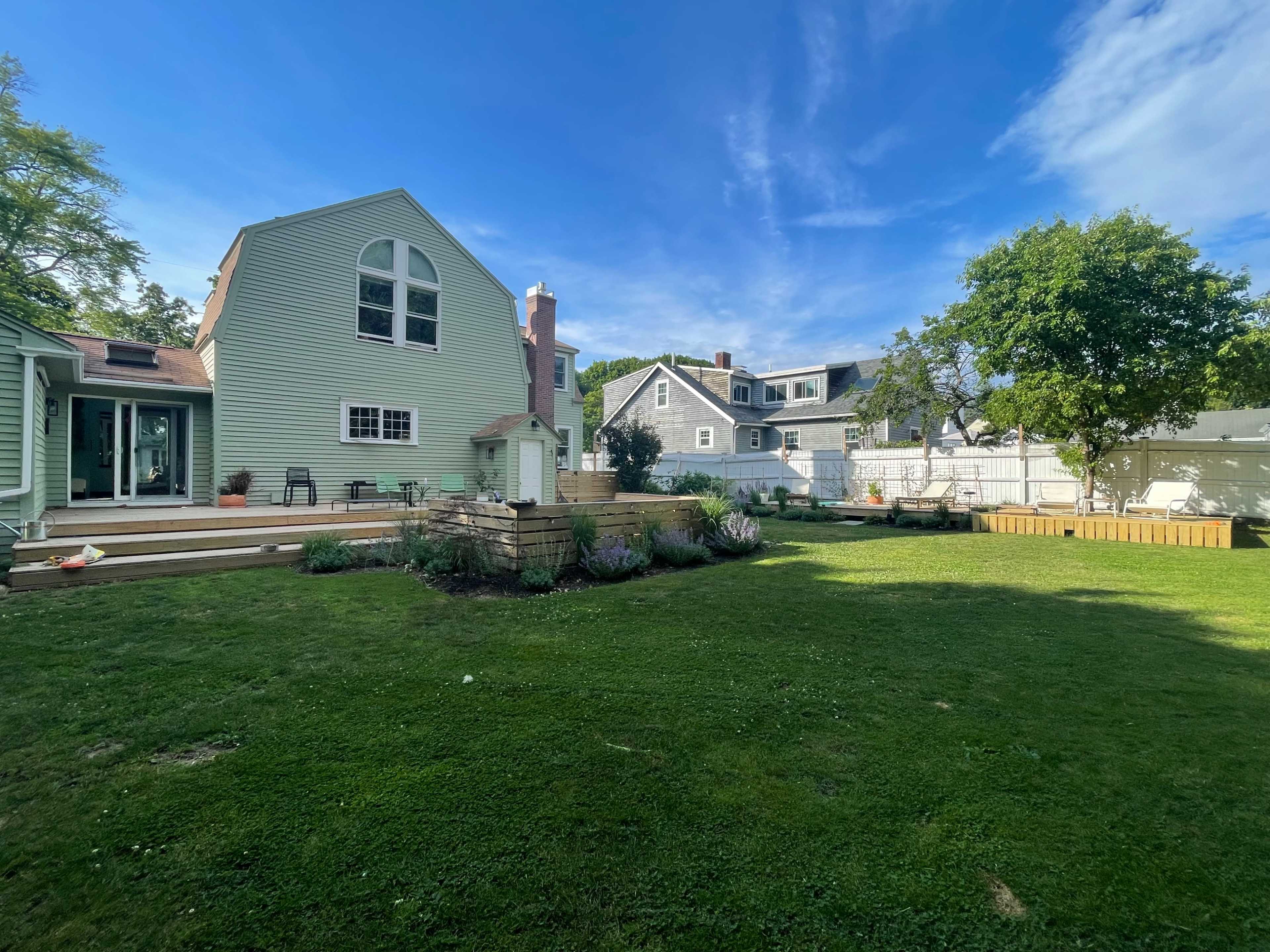 The image shows a spacious backyard with a green lawn, surrounded by multiple houses and trees under a clear blue sky.