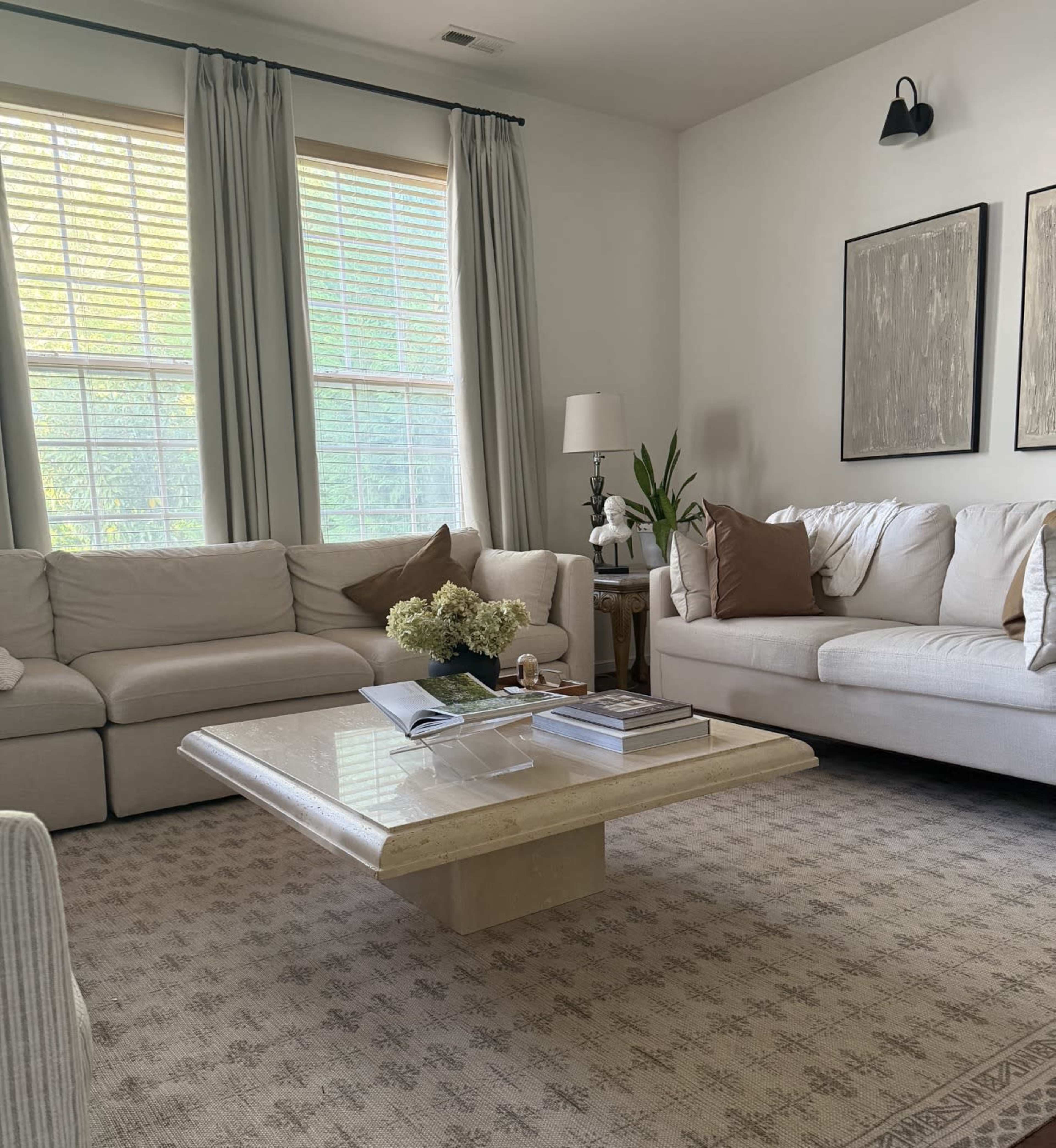 The image shows a cozy living room with two white sofas, a light-colored coffee table, and large windows adorned with curtains.