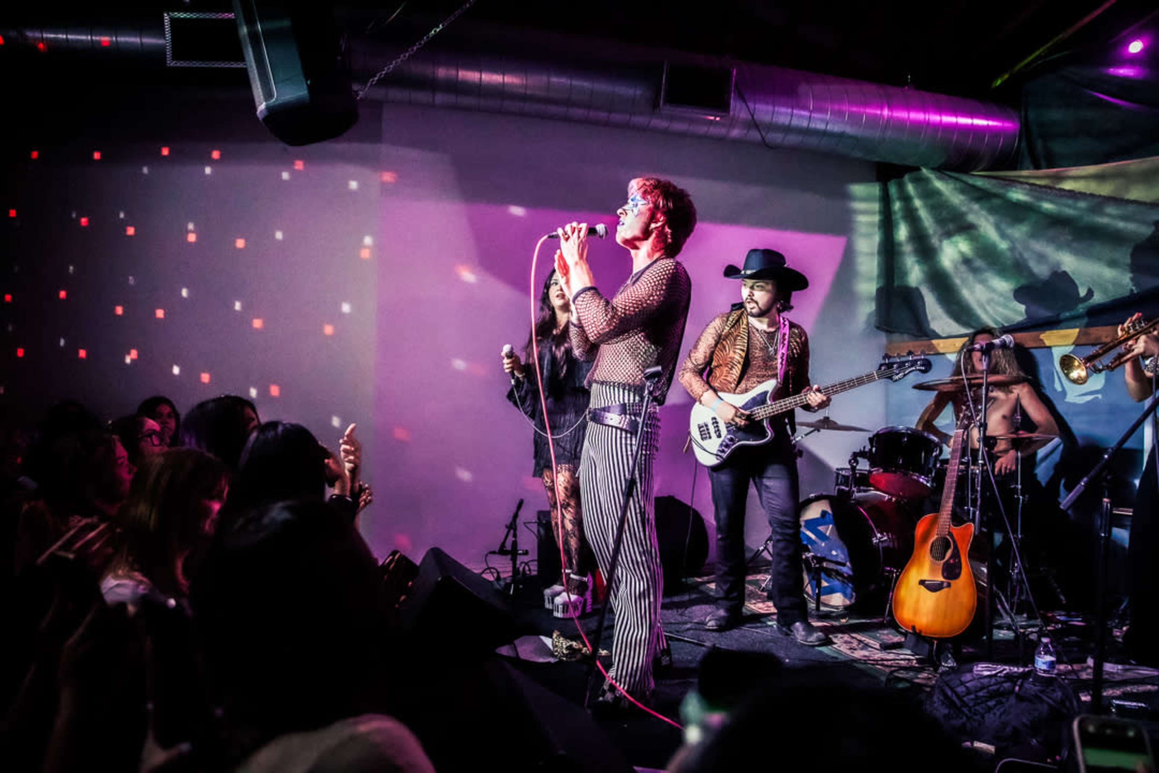 A singer performs energetically on stage while a band accompanies him, and an audience enjoys the live music.