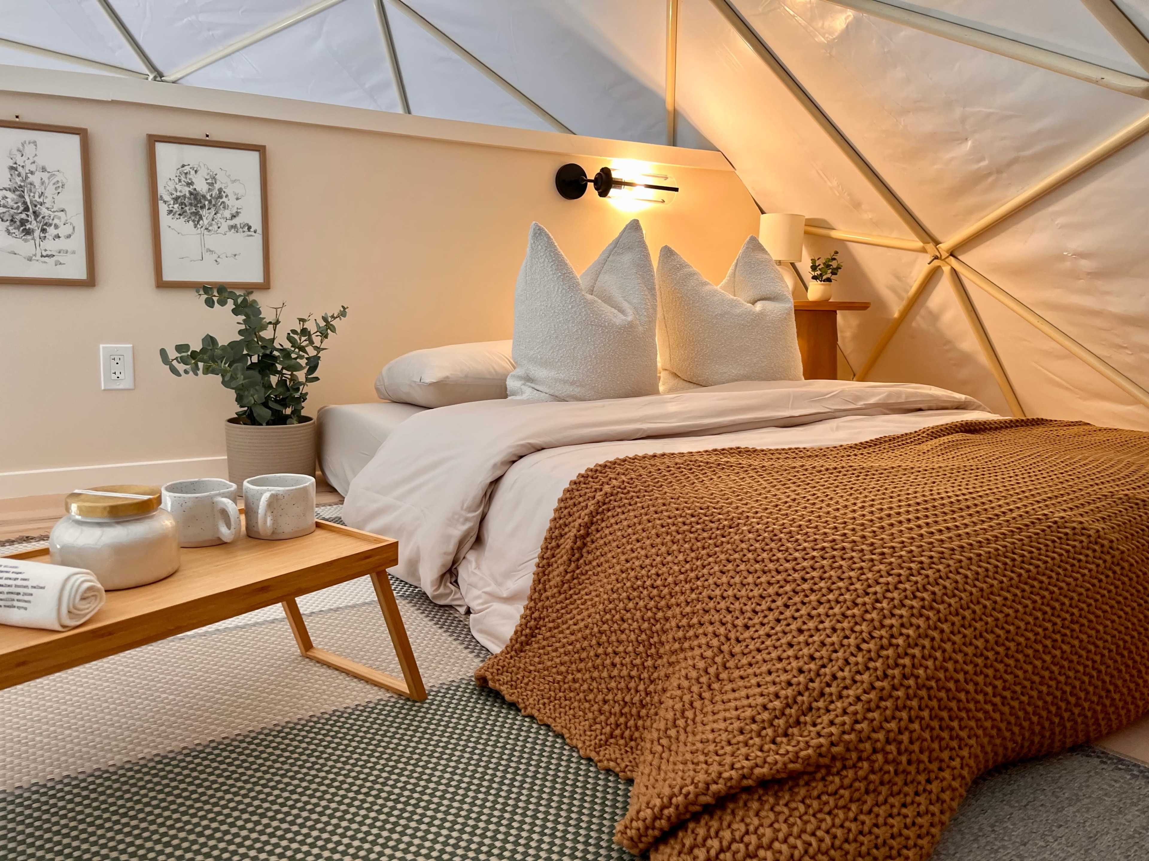 A cozy bedroom features a bed with large decorative pillows, a warm throw blanket, and a wooden table set with mugs and a towel.