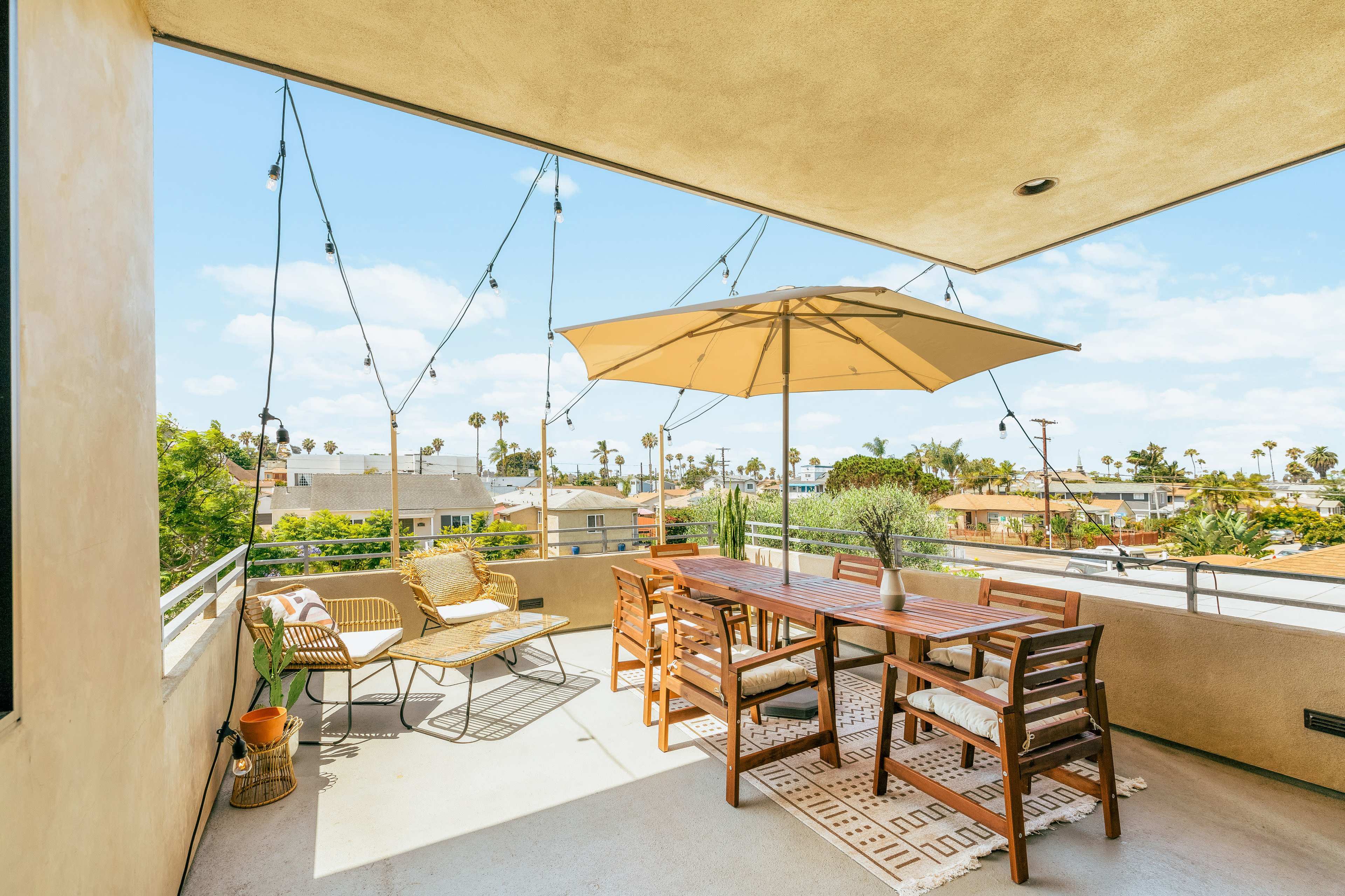 A spacious outdoor terrace features a dining table with an umbrella, string lights overhead, and seating arrangements with palm trees in the background.
