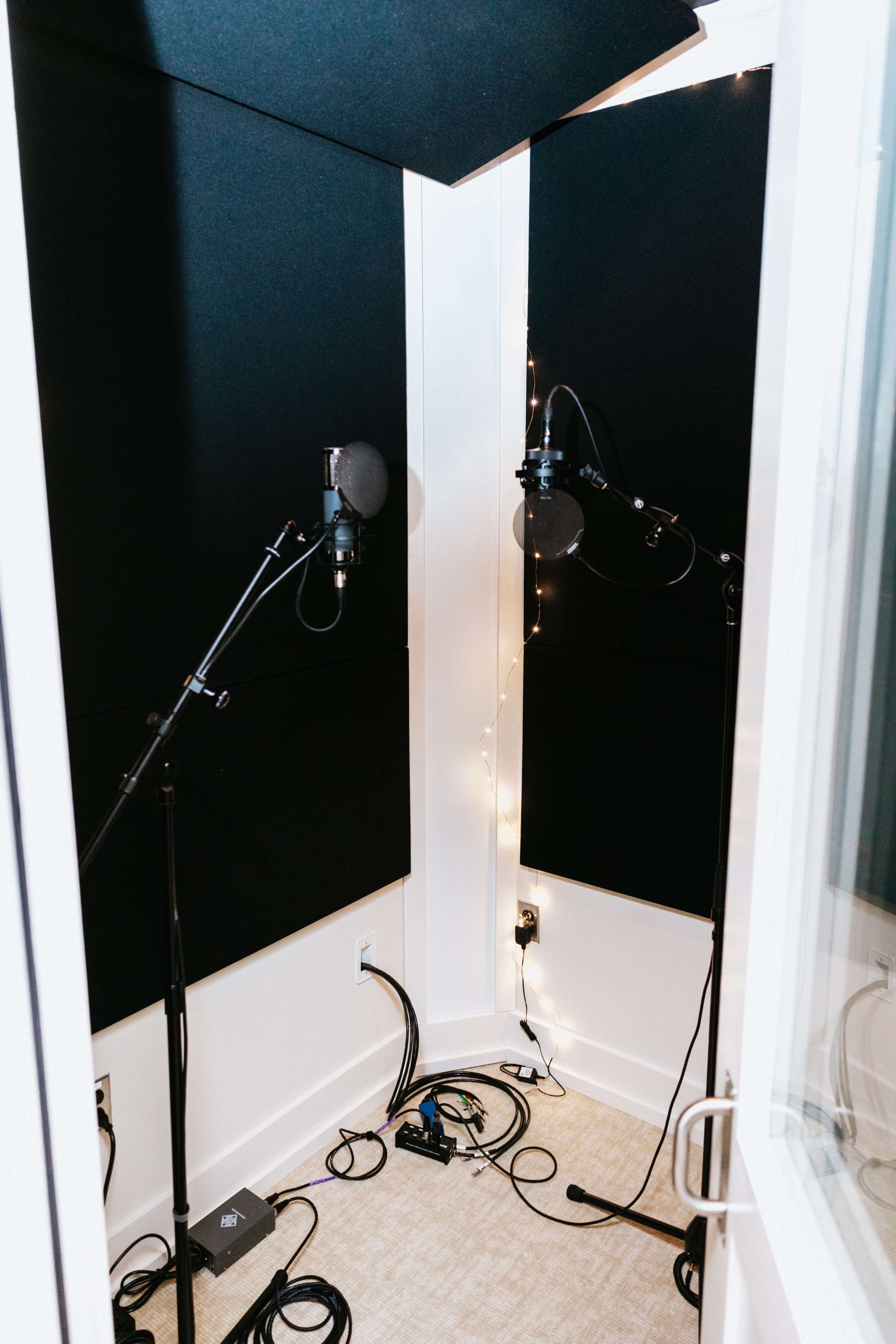 The image shows a recording booth with two microphones on adjustable stands and soundproofing panels lining the walls.
