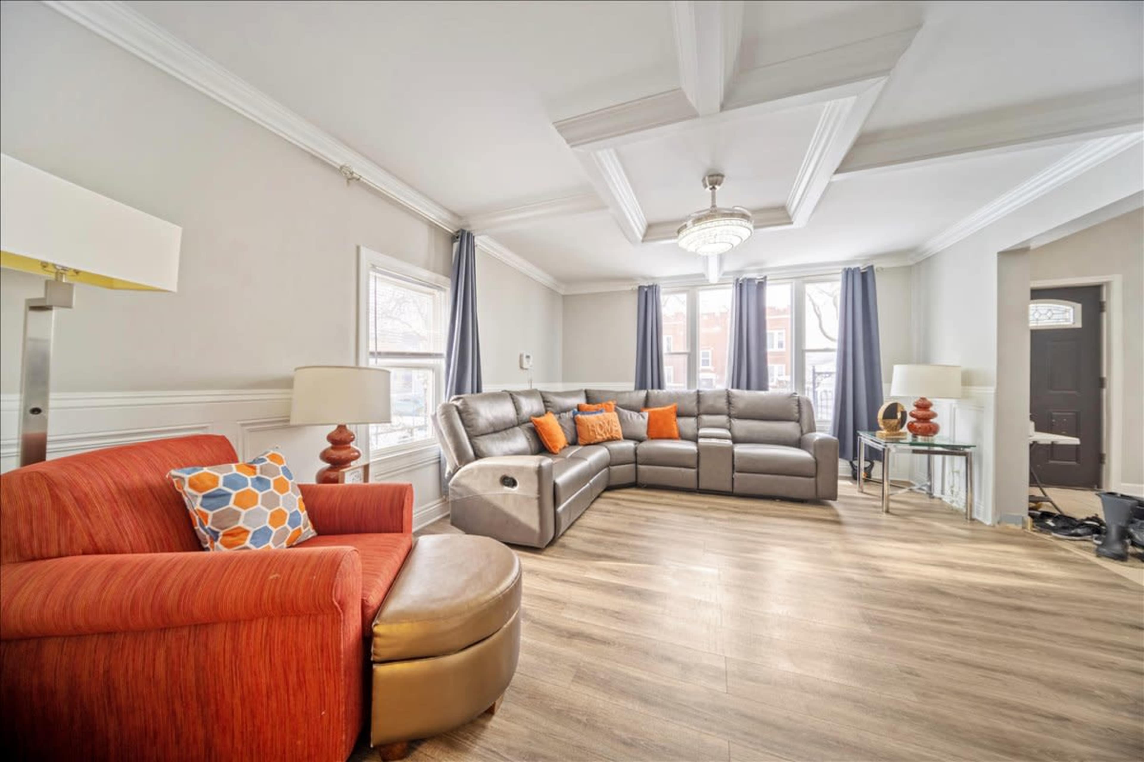 The image shows a spacious living room with a sectional sofa, an armchair, a large window, and hardwood flooring.