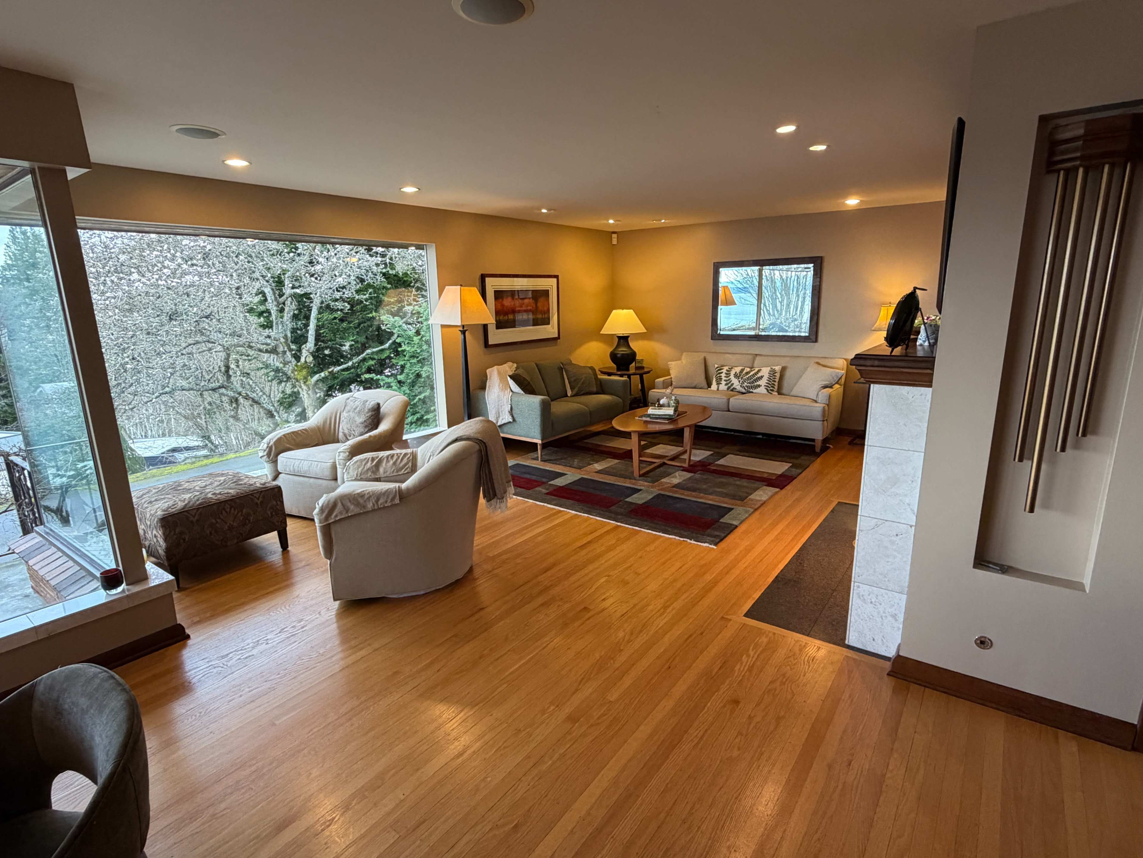 The living room features large windows overlooking a garden, with two sofas, a coffee table, and a couple of lamps arranged on a hardwood floor.