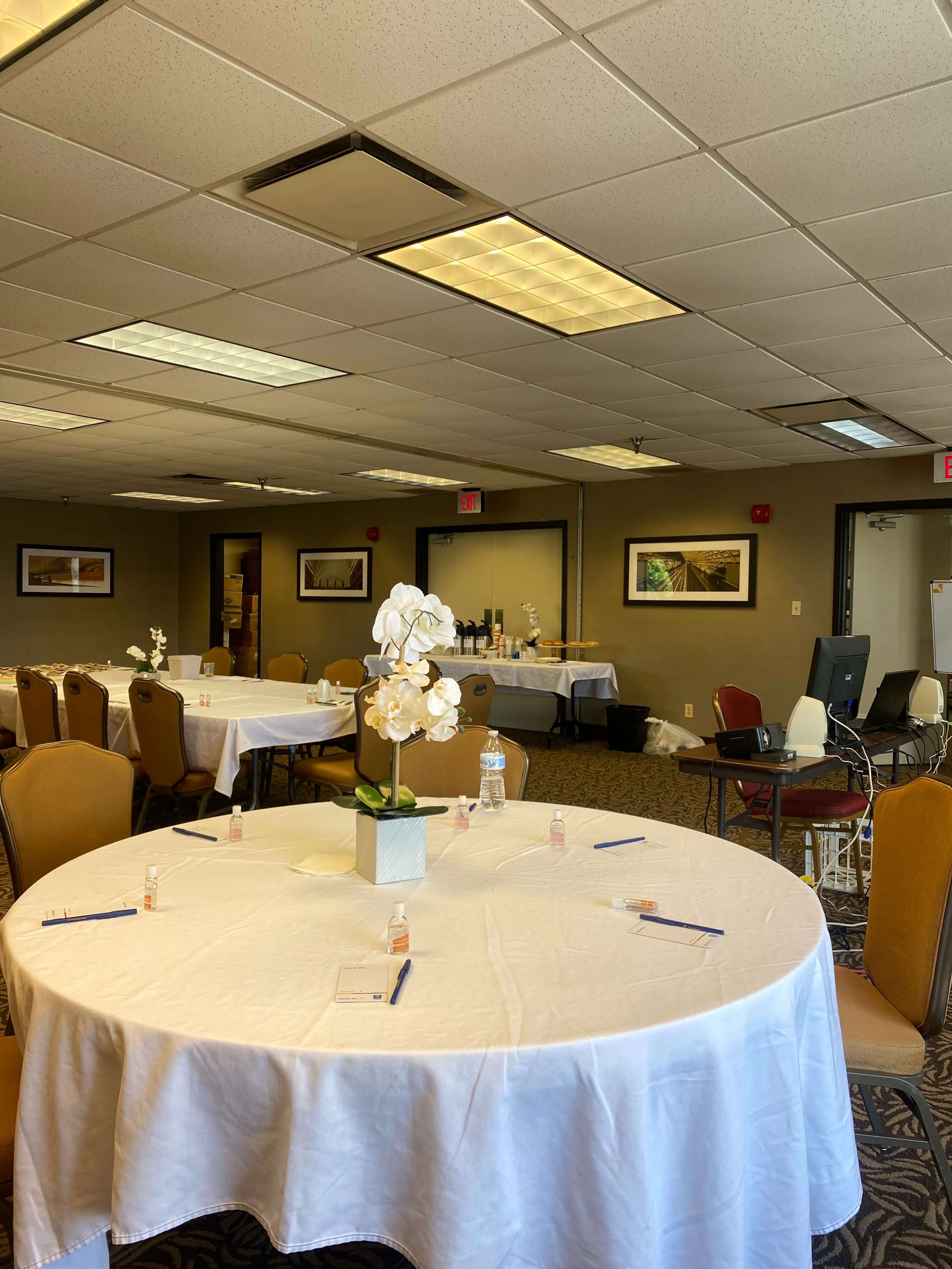 A conference room features several round tables covered in white tablecloths, each adorned with a small potted orchid and writing materials, while the walls display framed artwork.