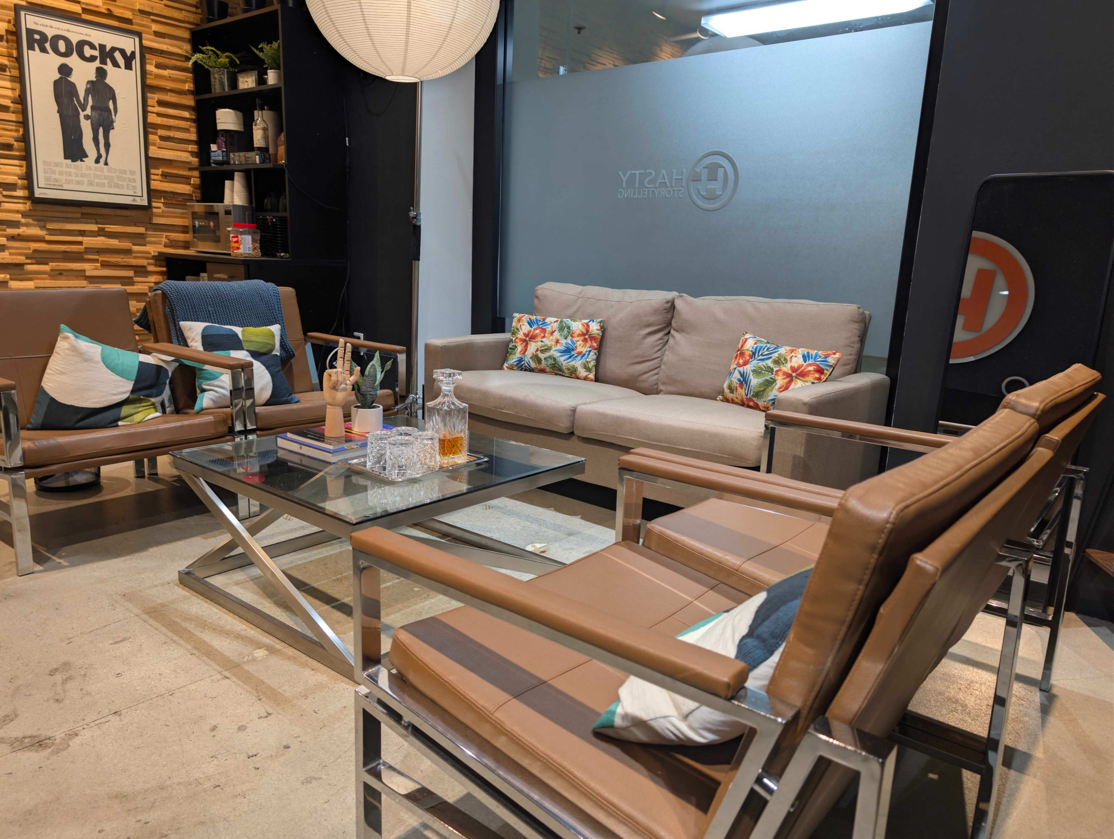 The image shows a cozy seating area with a sofa, armchairs, and a glass coffee table, surrounded by wooden decor and a backdrop featuring a logo.