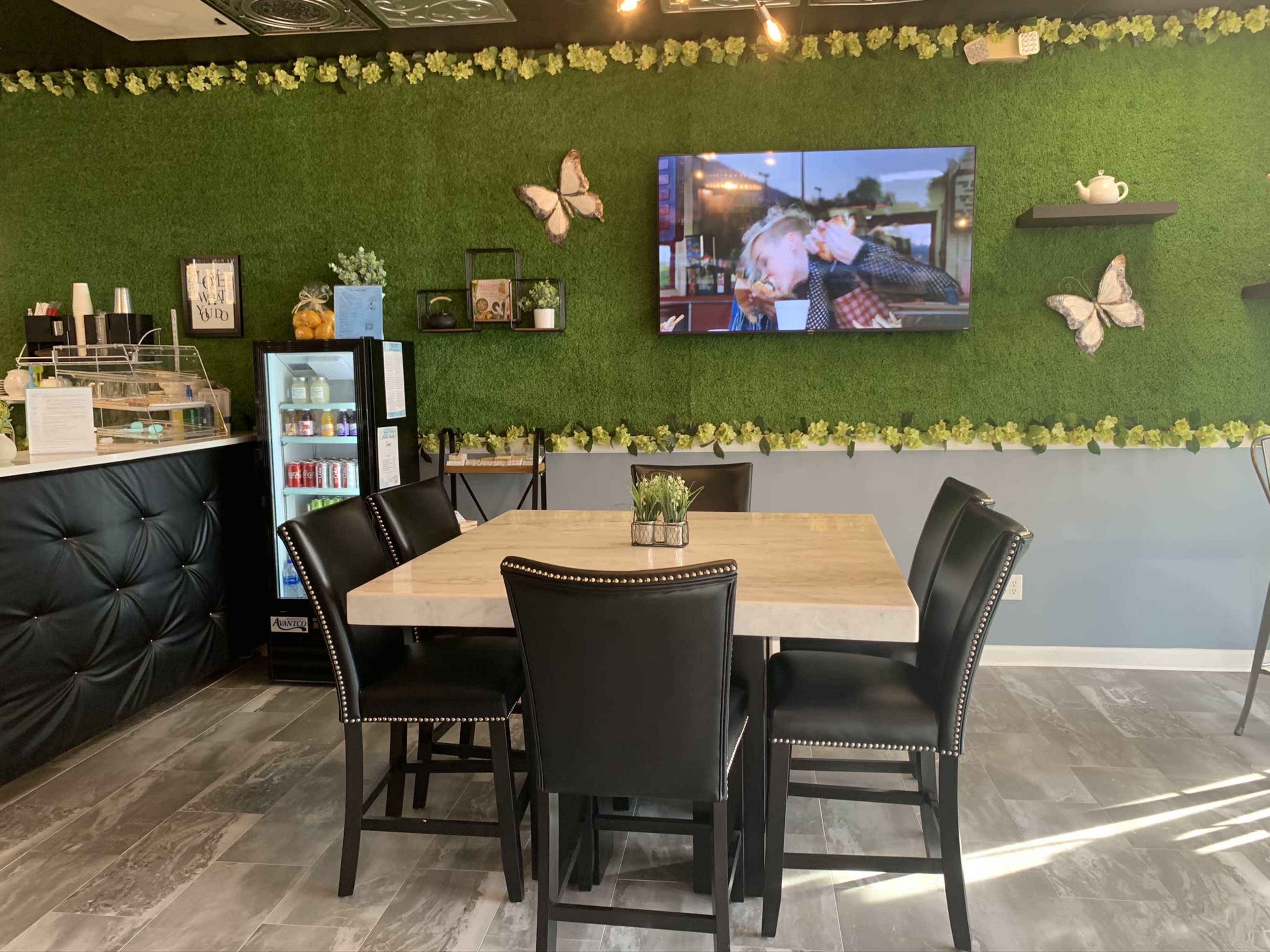 A dining area features a wooden table surrounded by black chairs, with a green wall adorned with artificial plants and a television displaying a video.