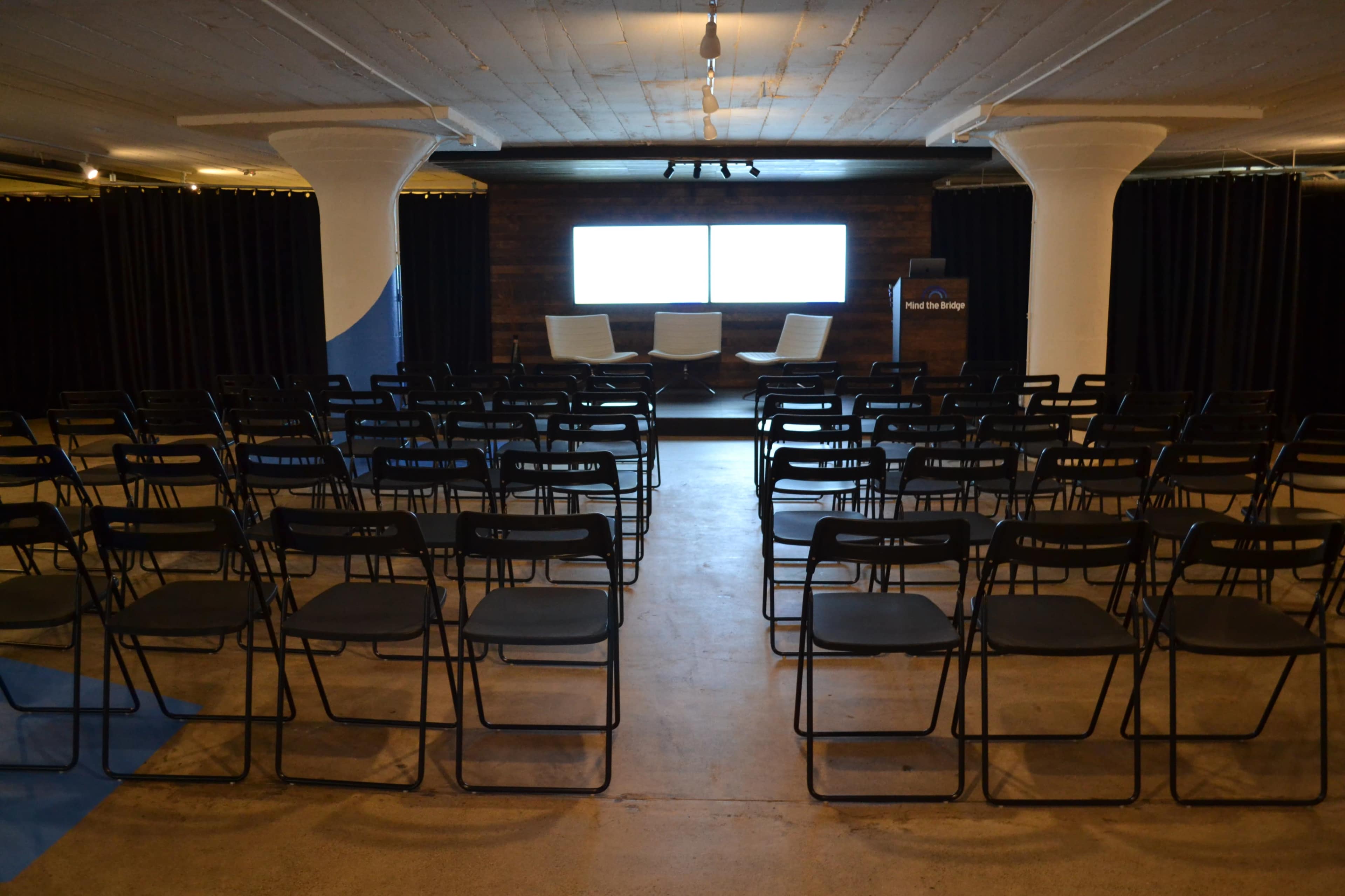 A setup for an event with neatly arranged black chairs facing a small stage featuring three white chairs and a large screen in a dimly lit room.