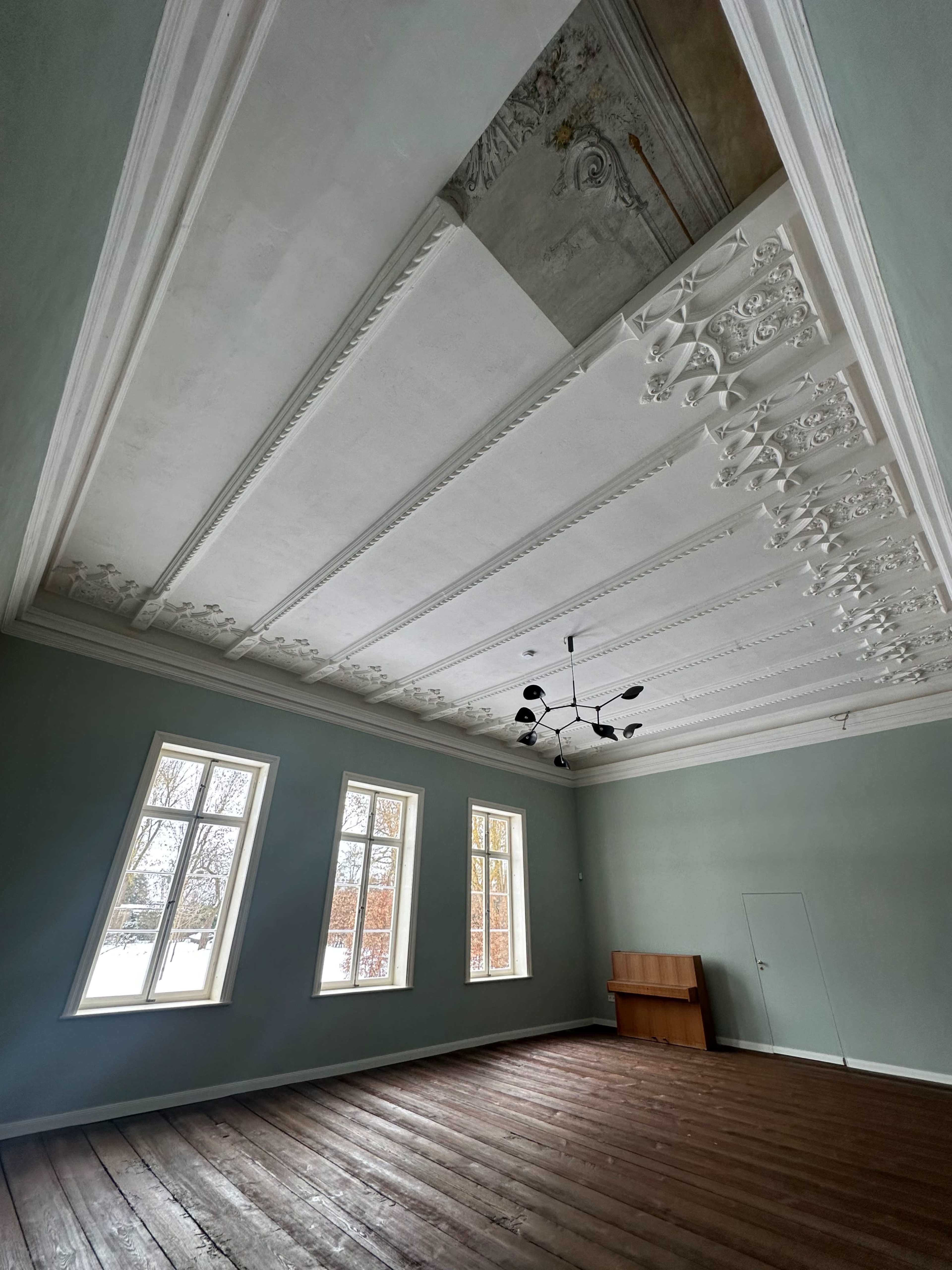 The image shows a spacious room with a wooden floor, high white ceiling featuring decorative molding, and large windows allowing natural light to enter.