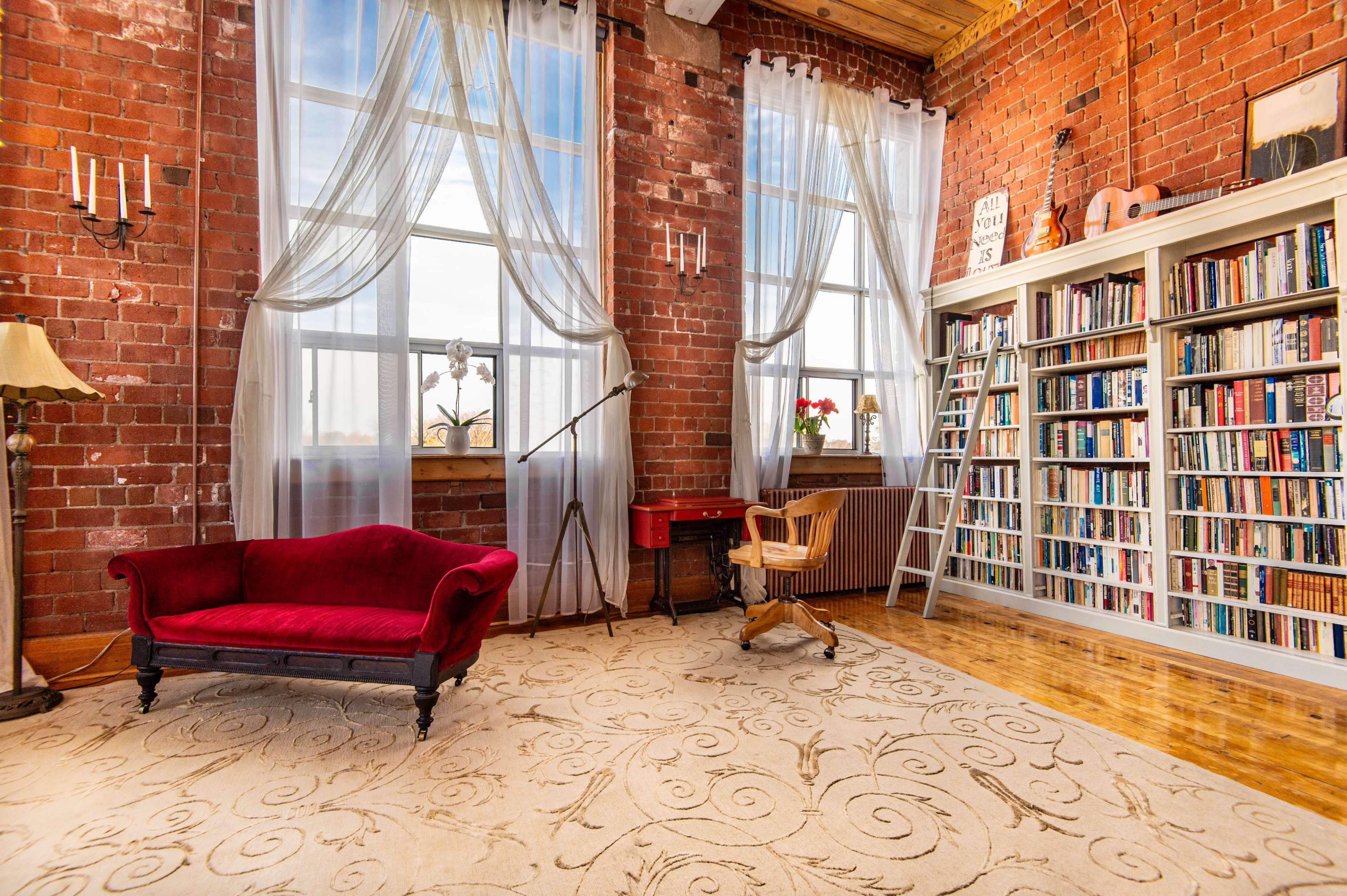 A cozy reading room with exposed brick walls, a large bookshelf filled with books, a red velvet couch, and a wooden chair positioned on a decorative rug.