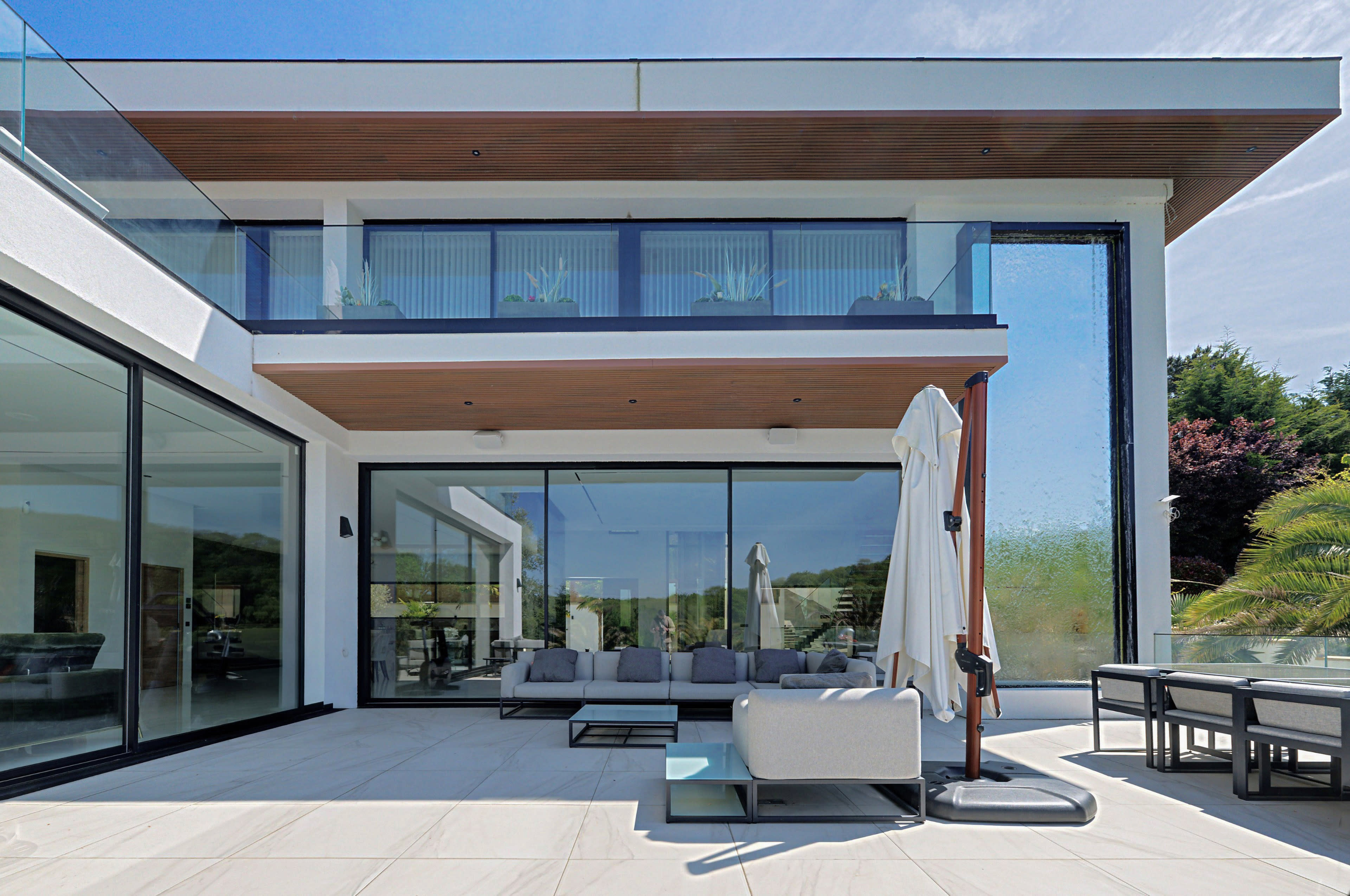 The image shows a modern two-story house with large glass windows, a patio area furnished with seating, and a stylish outdoor dining space.