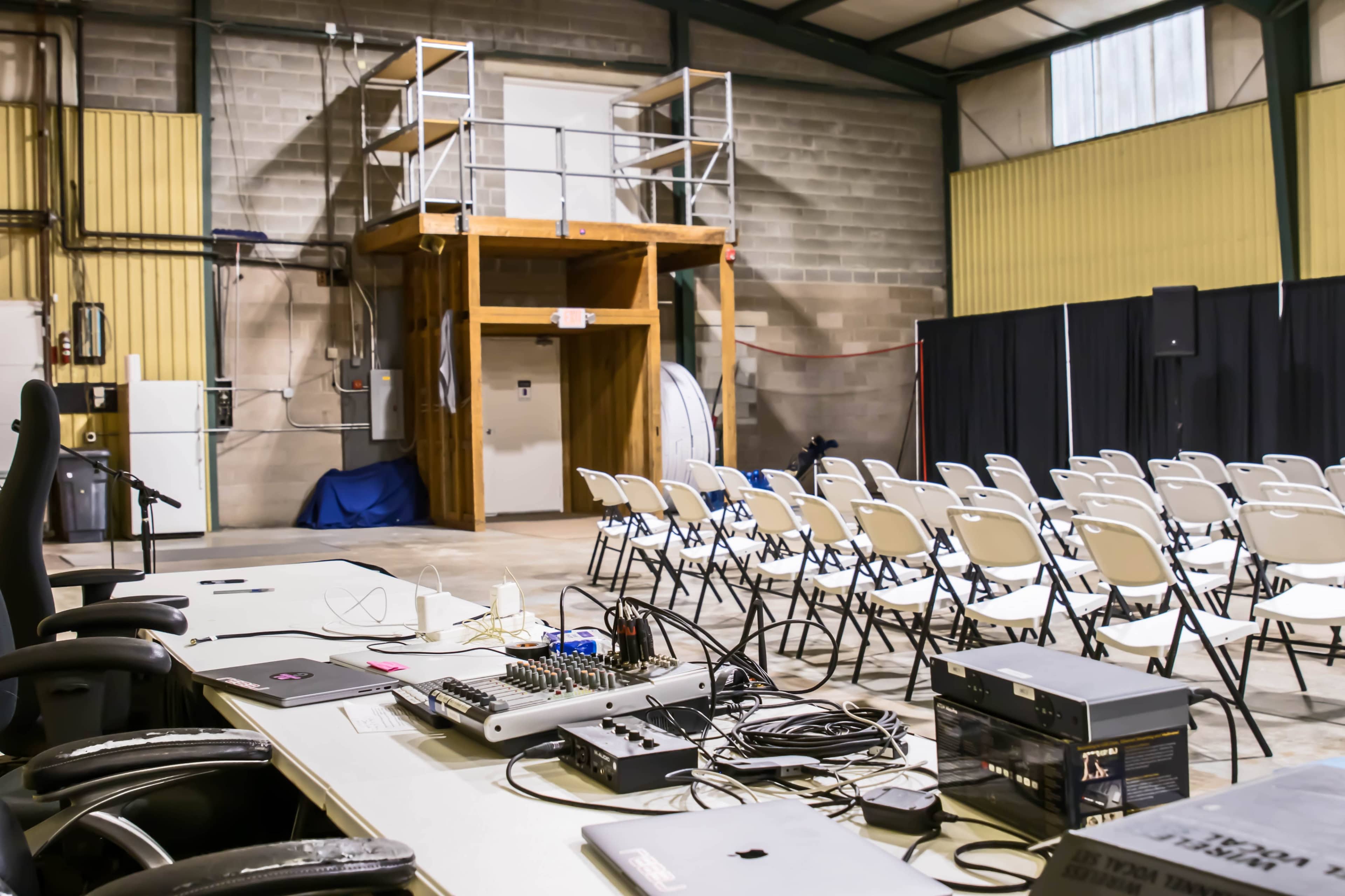 A large indoor space with rows of chairs set up facing a wooden structure and a sound mixing table with various cables and equipment on a table.