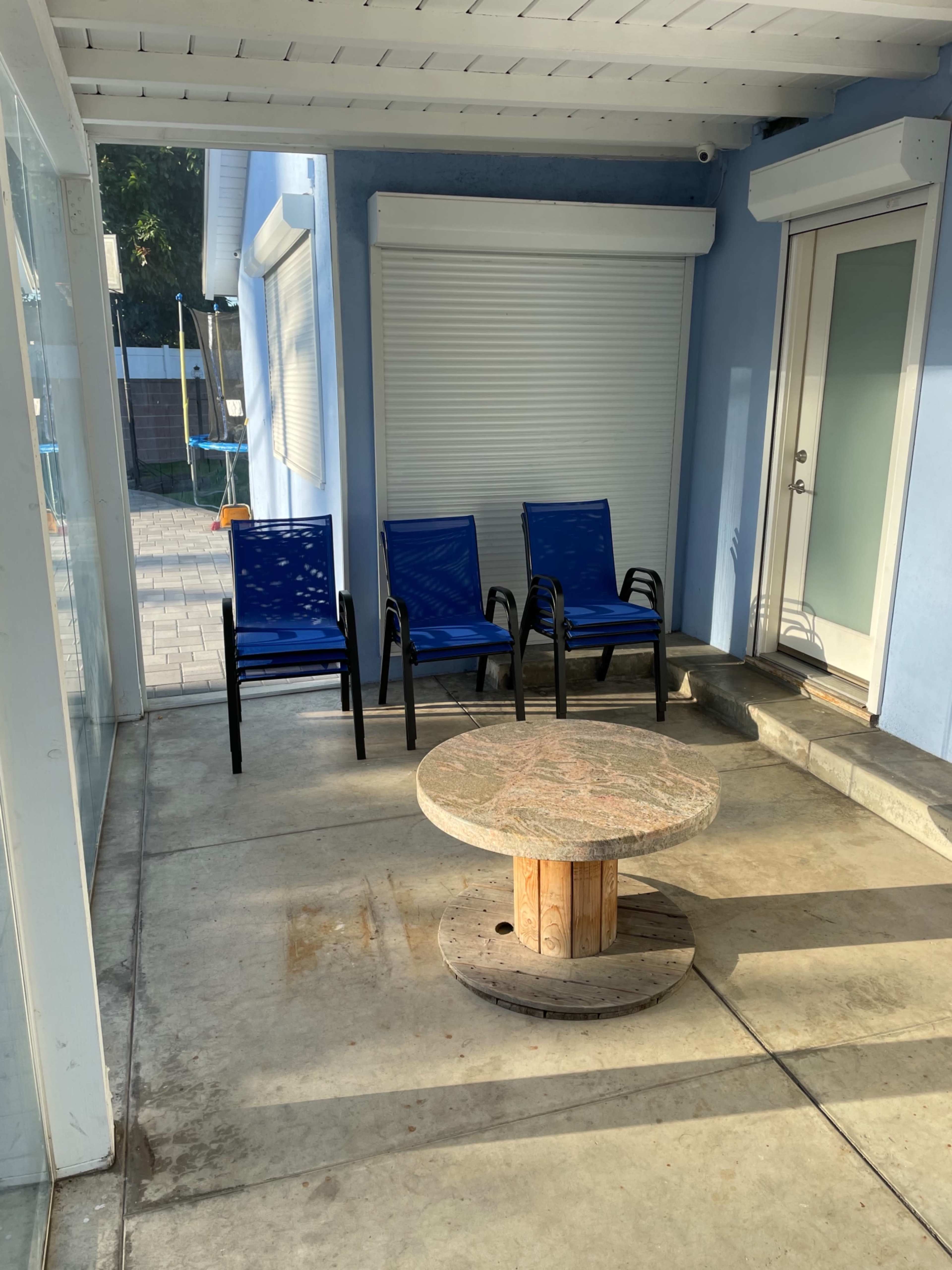 A covered patio area with three blue chairs and a circular wooden table on a concrete floor.