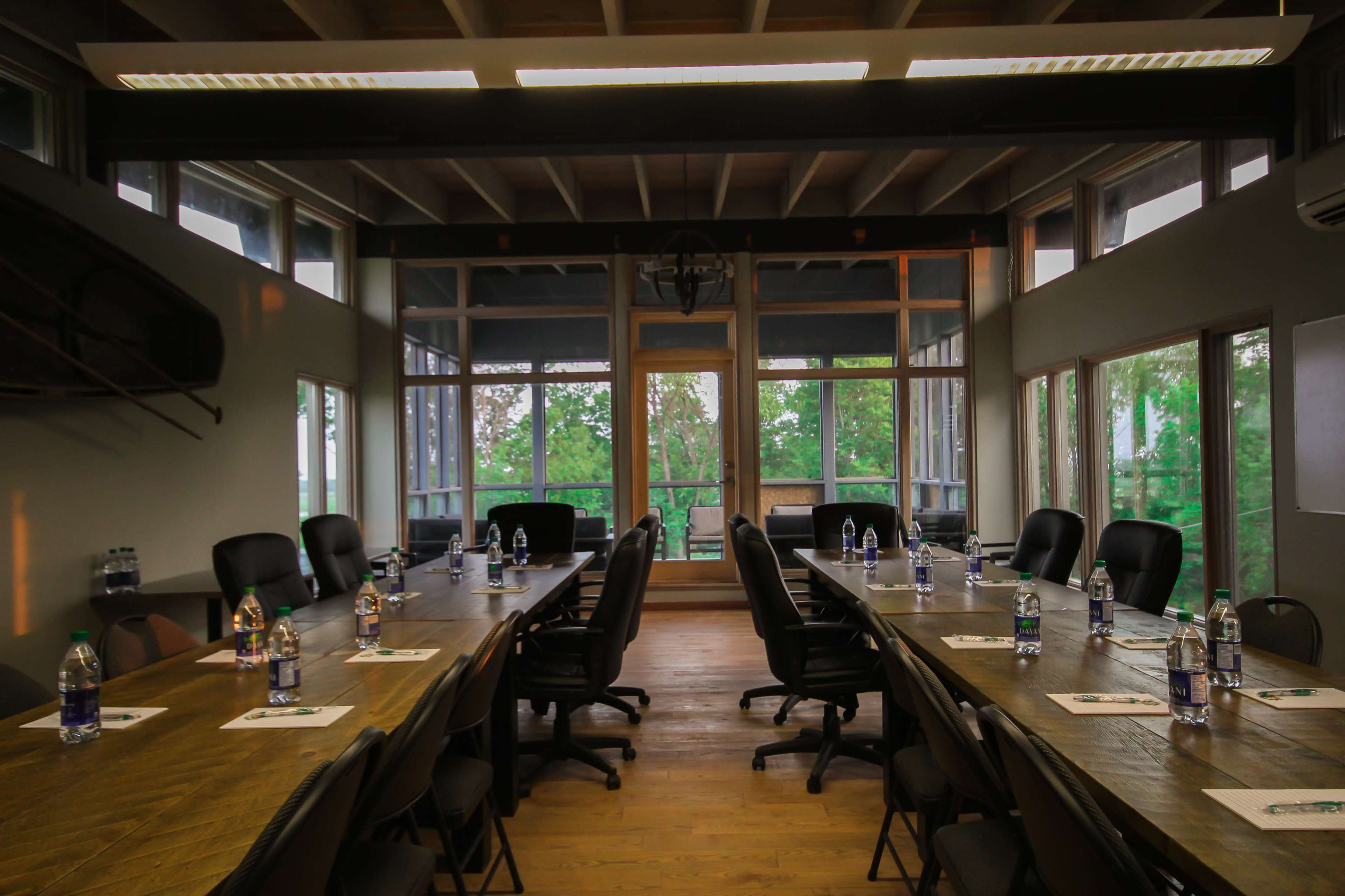 The image shows a conference room with two long tables arranged in a U-shape, set with water bottles and notepads, surrounded by chairs and large windows overlooking greenery.