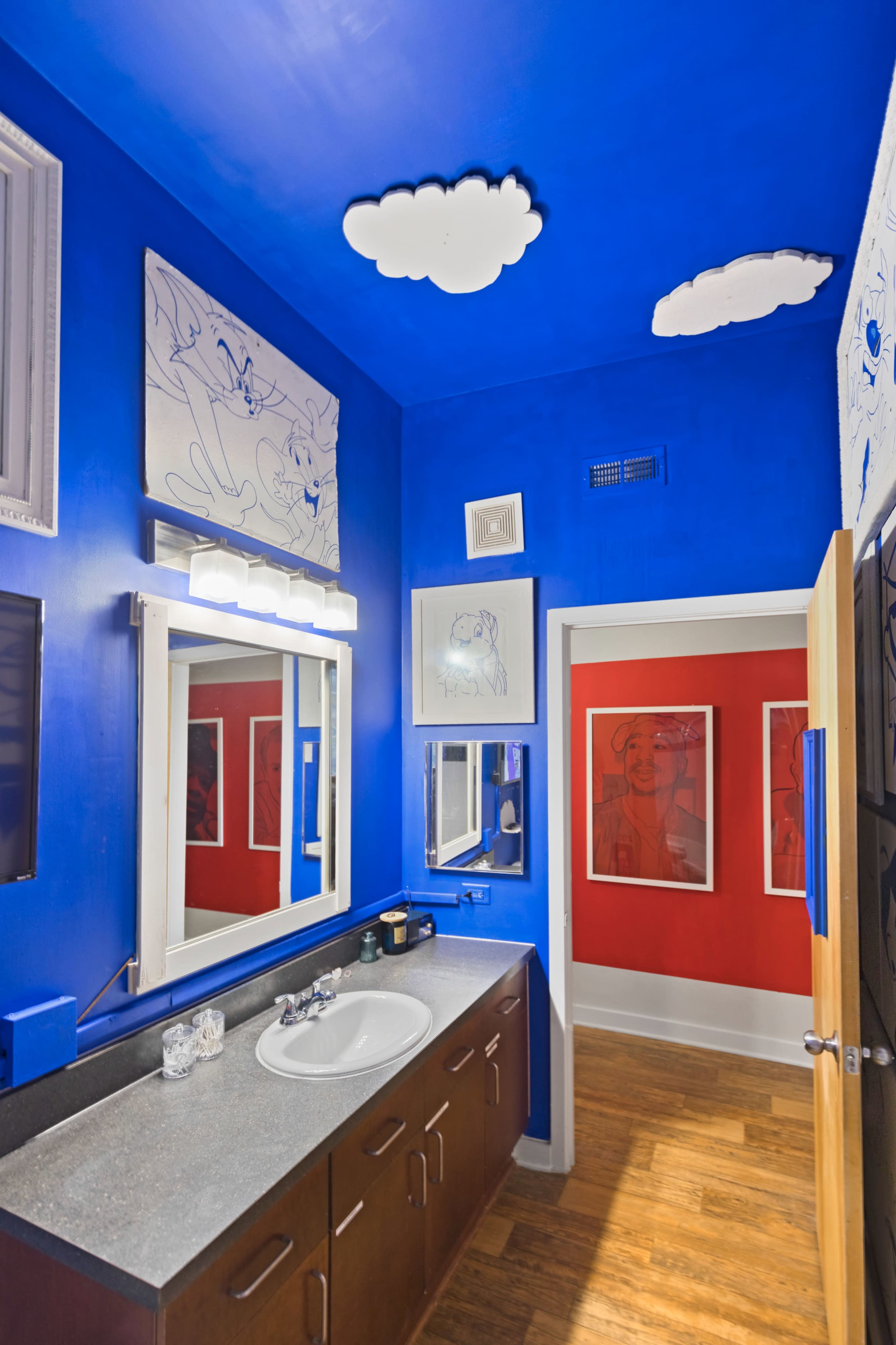 A brightly painted blue bathroom with a countertop sink, framed artwork on the walls, and a doorway leading to a red-walled room.