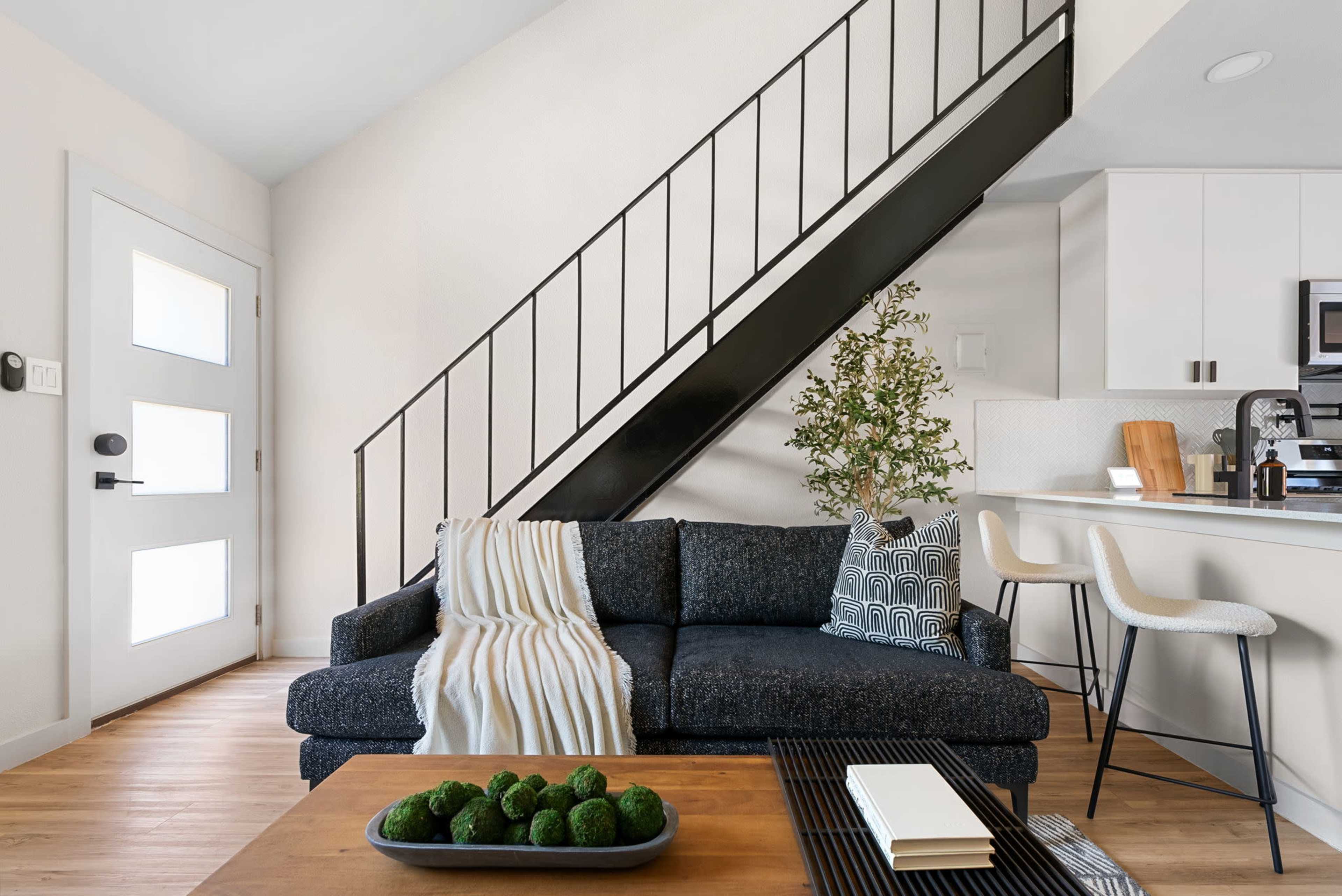 The image shows a modern living space featuring a dark couch with a decorative pillow, a wooden coffee table with green decor, and a staircase leading to an upper level.