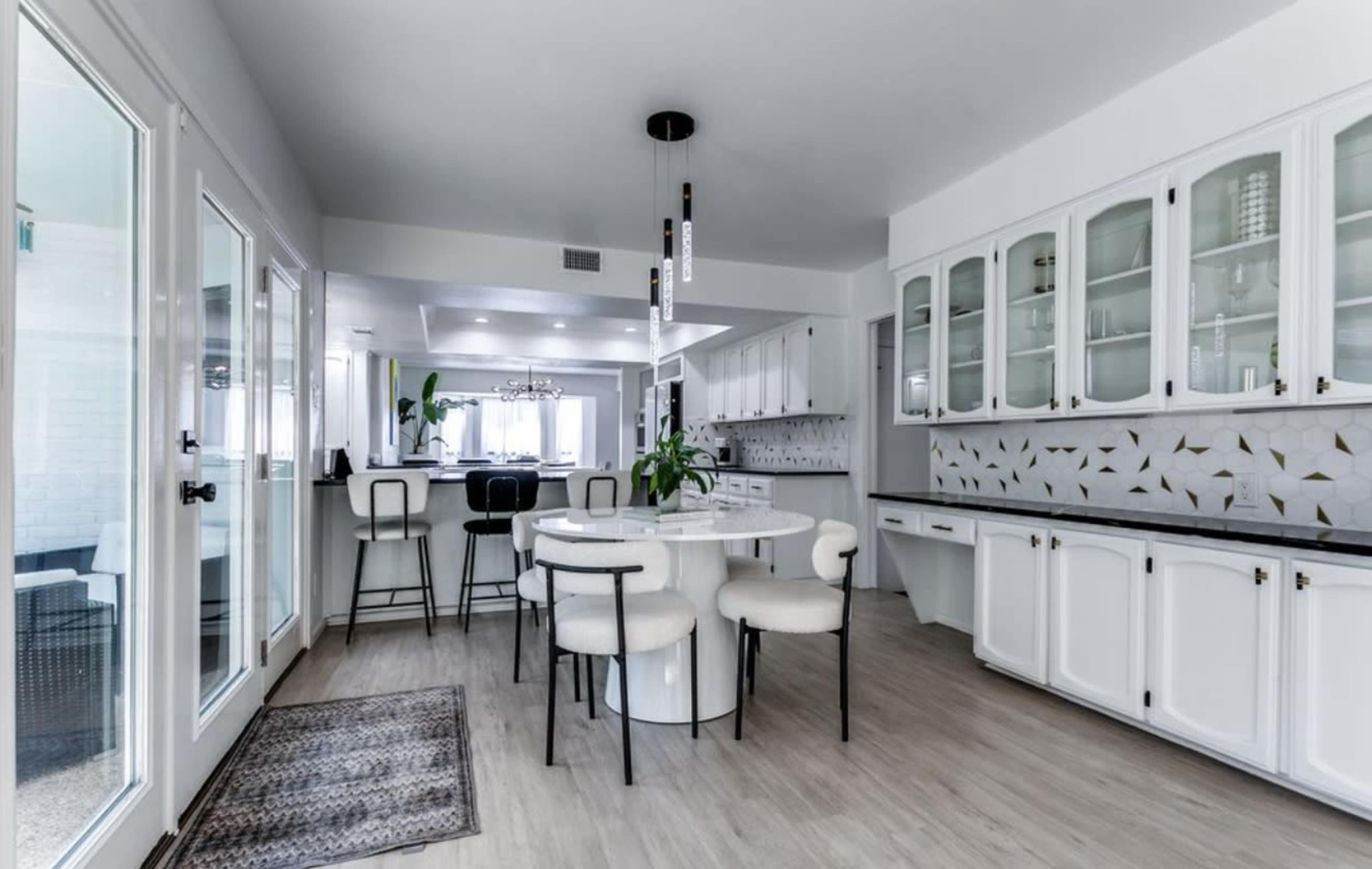 The image shows a modern kitchen with white cabinets, a round dining table, and a bar area with high stools.