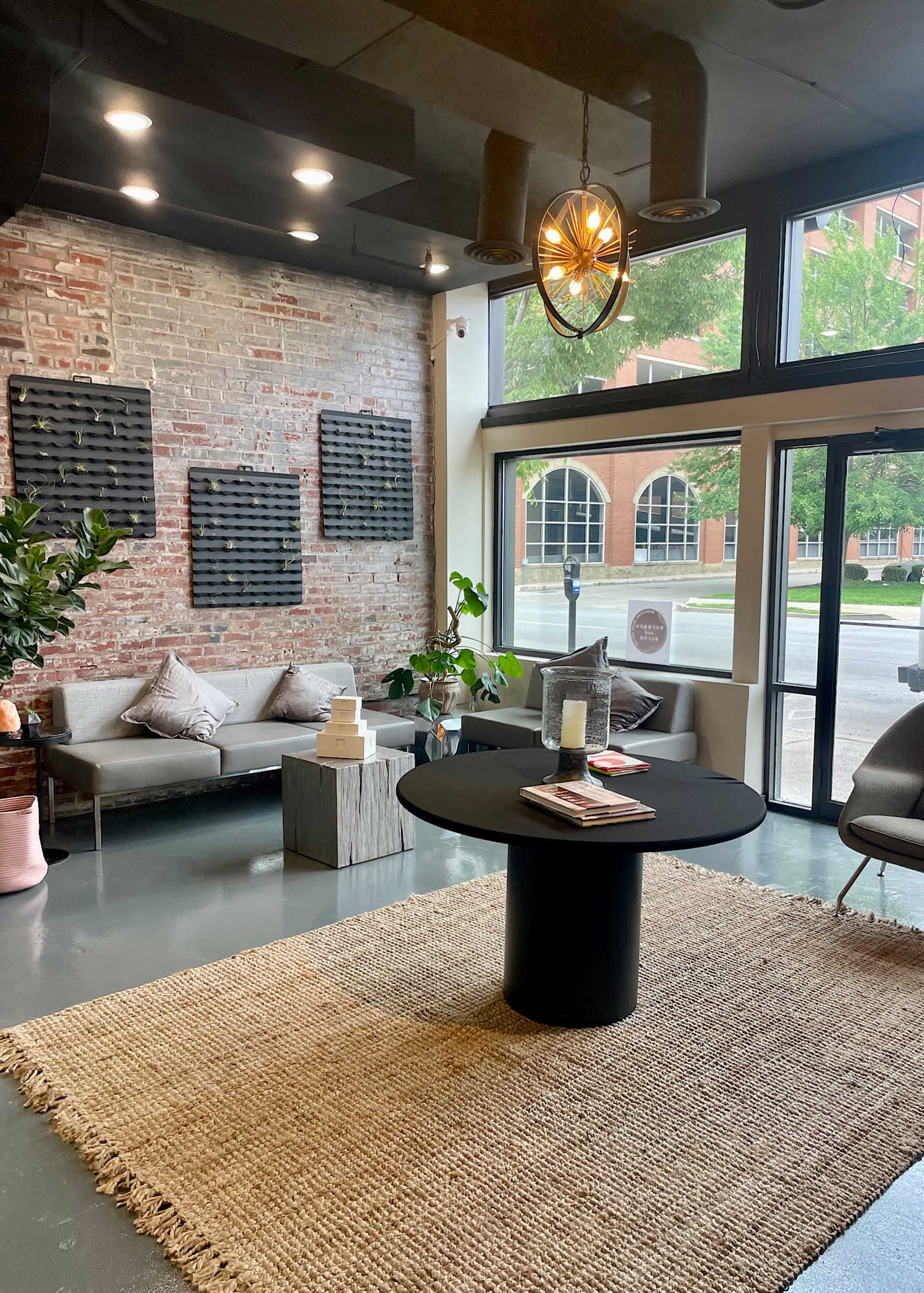 A modern indoor space featuring a seating area with a gray sofa, a round black table, and large windows facing a street, complemented by a woven rug and plants.