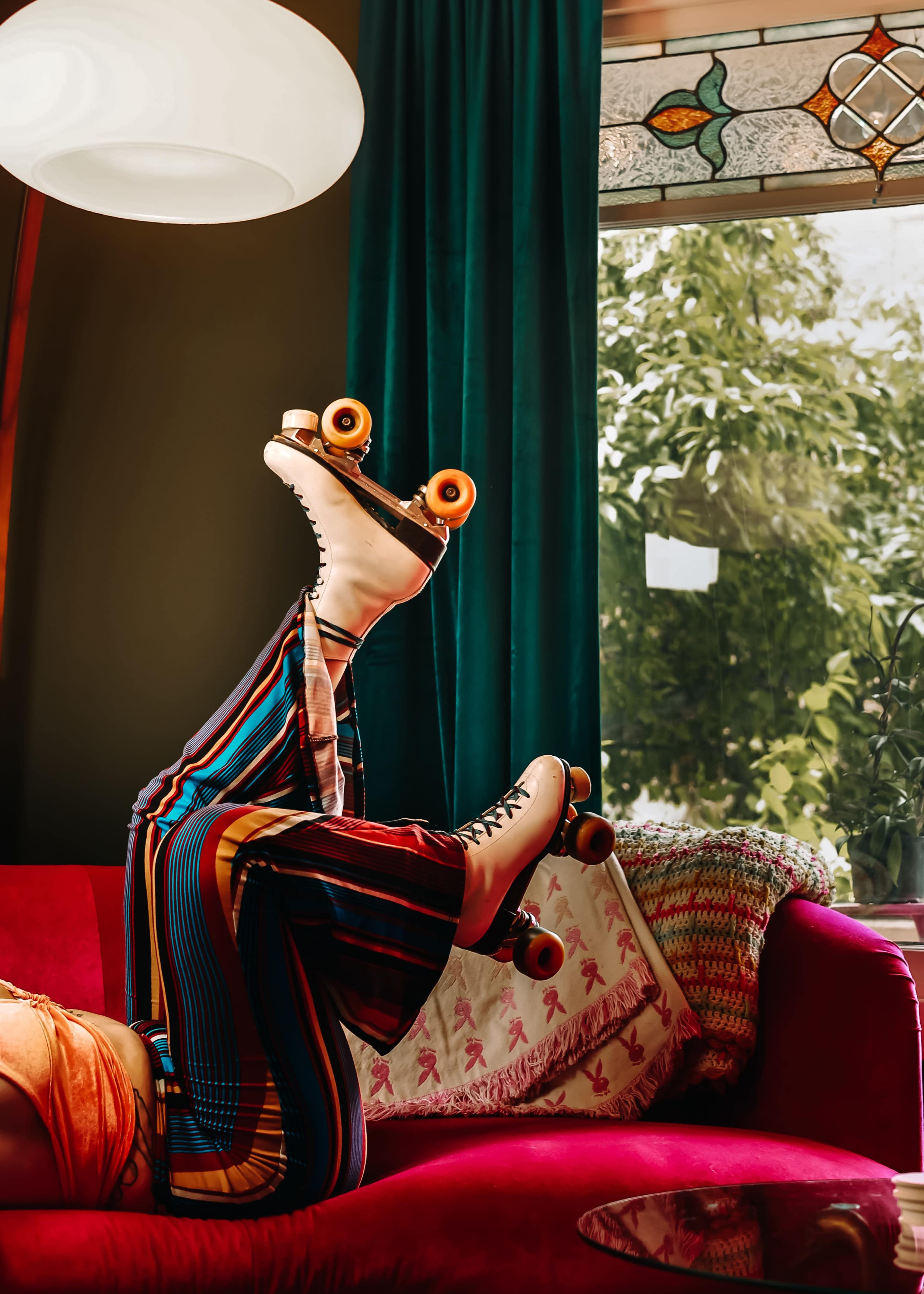 A person in colorful striped pants is lying upside down on a red couch, wearing roller skates.