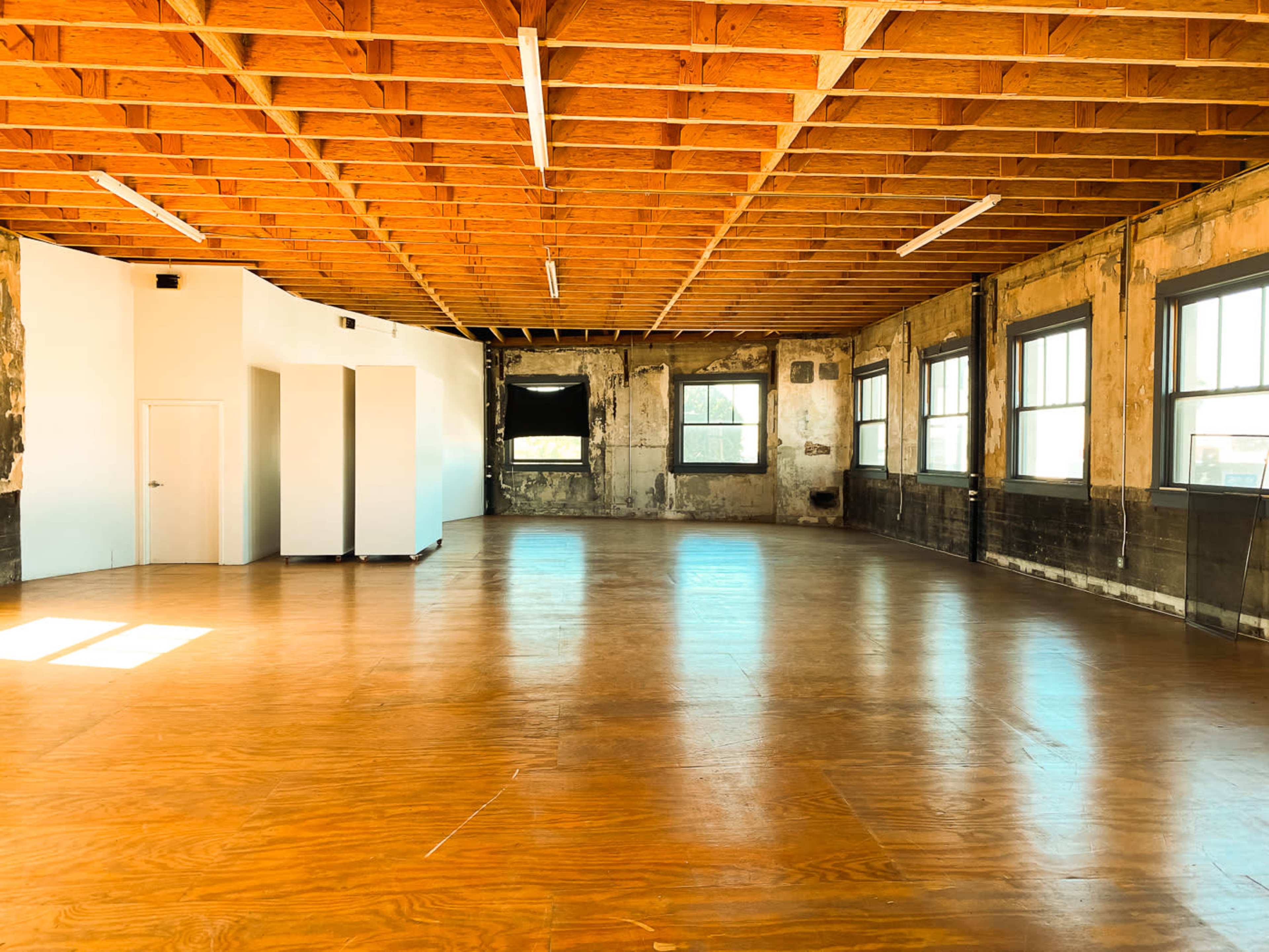 The image shows a spacious, empty room with wooden flooring, exposed beams, and large windows.