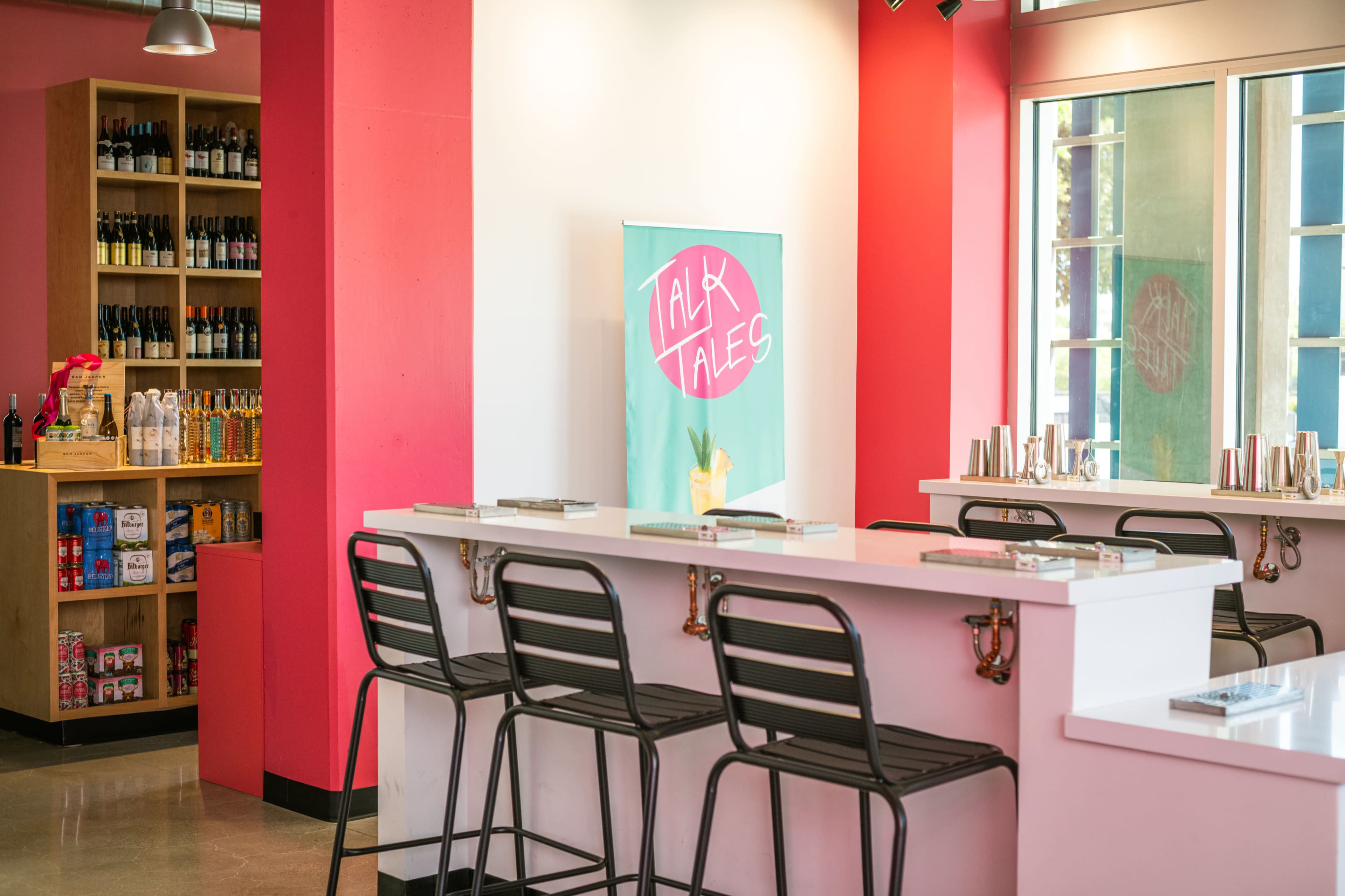 The image shows a modern bar area with a white countertop, black stools, and a display of bottles and products on a wooden shelf in the background.