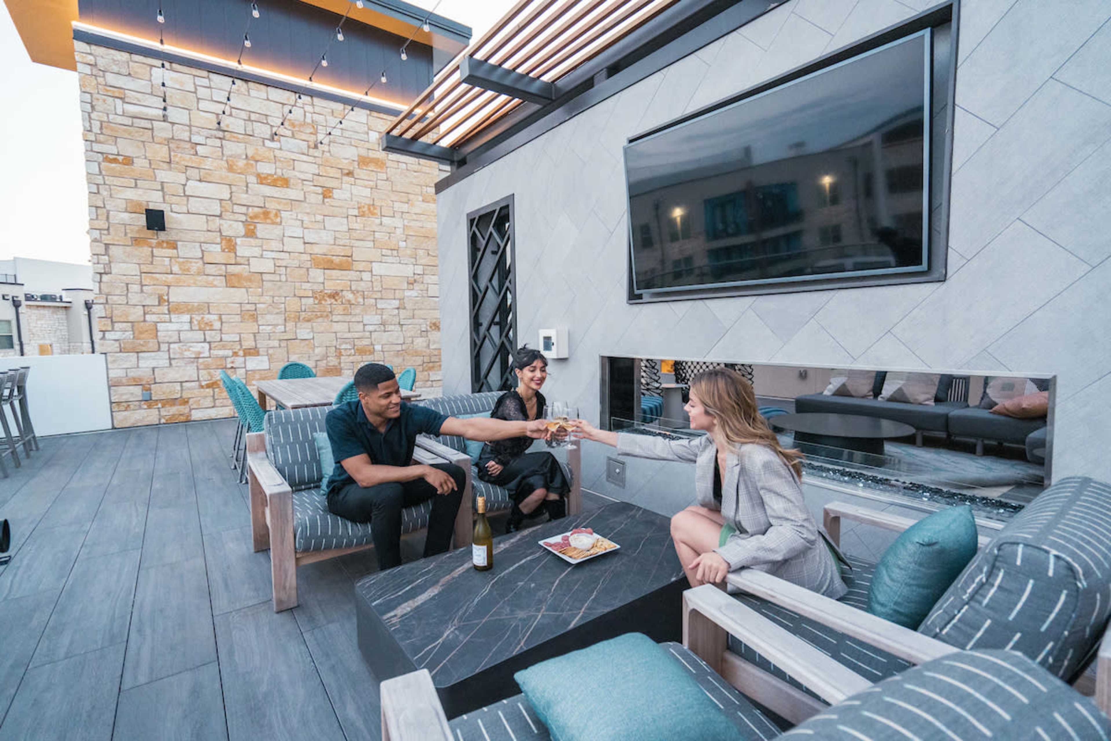 Three people sit on a modern rooftop lounge, toasting with glasses of champagne beside a table with a plate of food.