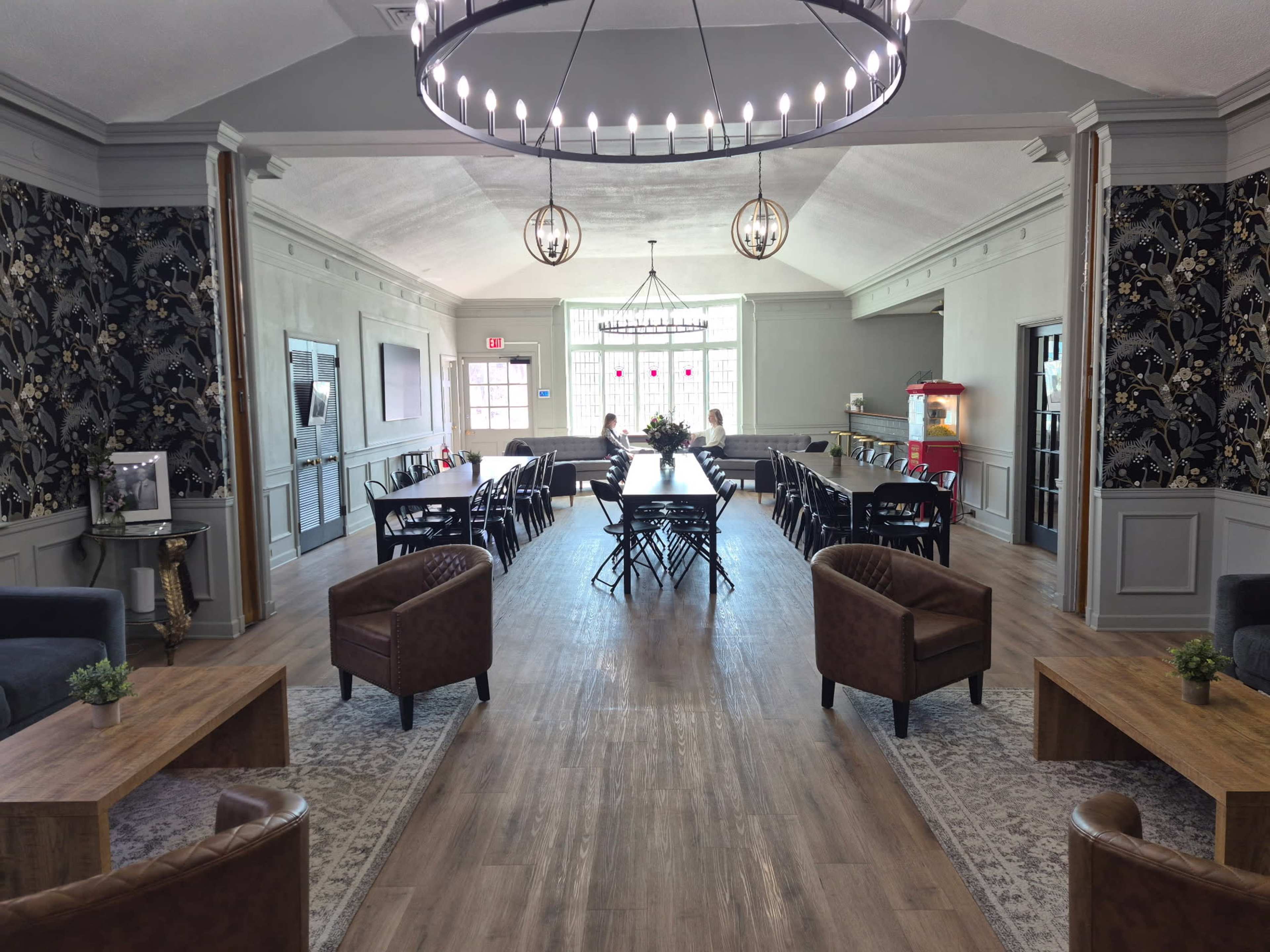 The image shows a spacious, elegantly decorated room with a long dining table surrounded by chairs, potted plants, and a seating area featuring brown leather chairs and tables.