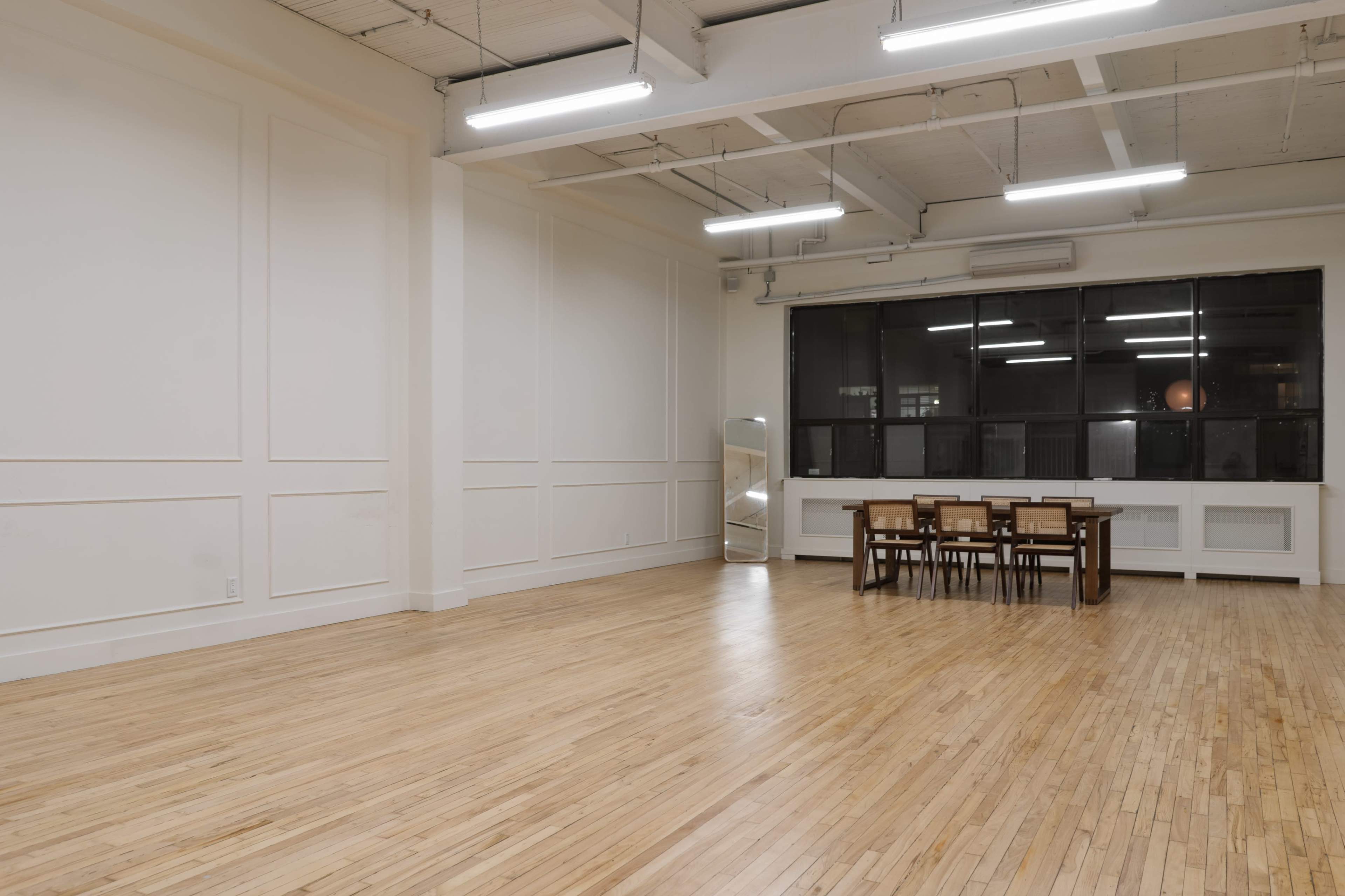 A spacious room with wooden flooring features a large window and a set of four wooden chairs around a dining table.