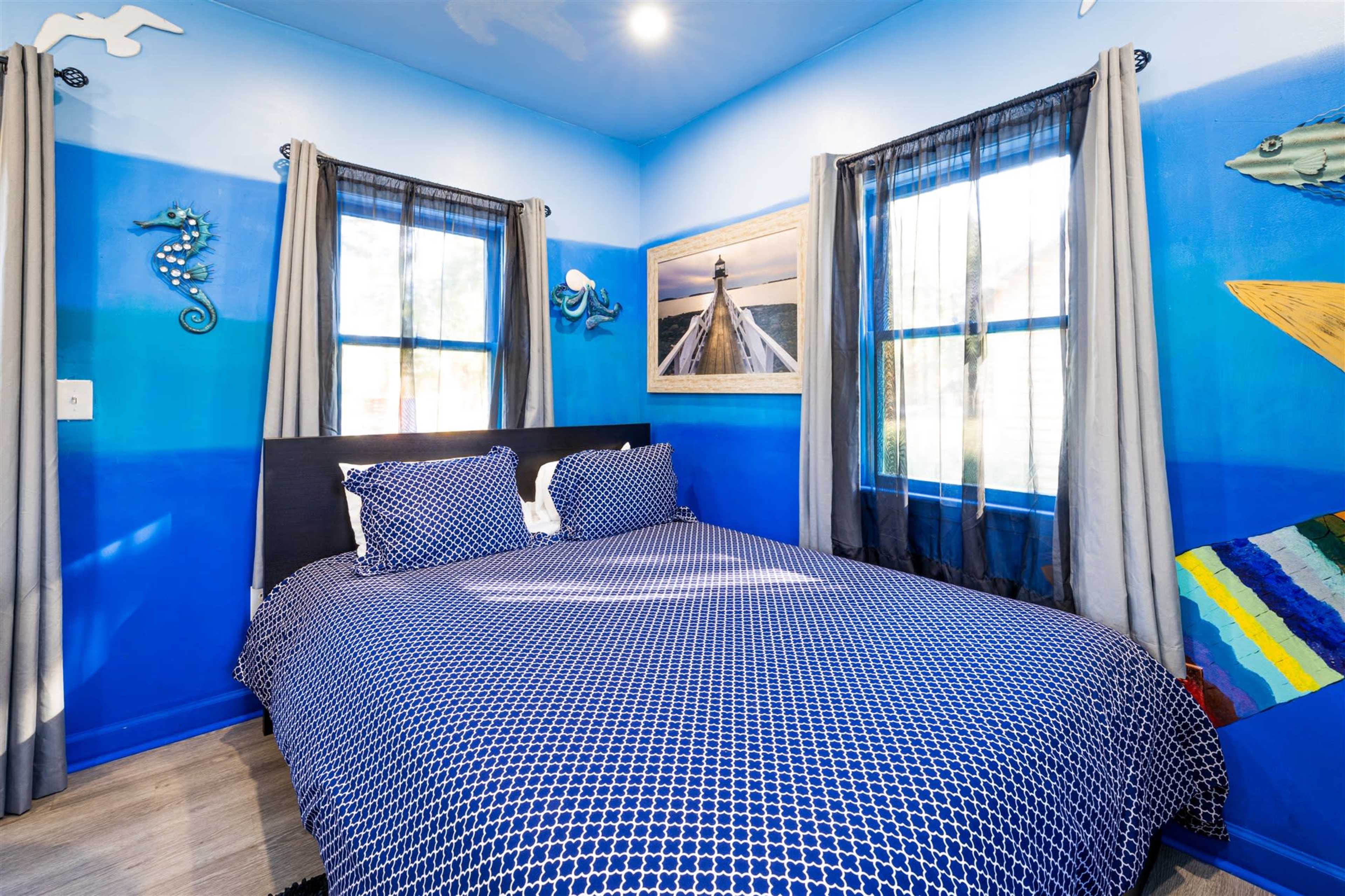 A brightly painted bedroom features blue walls with marine-themed murals, a bed with a patterned duvet cover, and large windows covered by gray curtains.