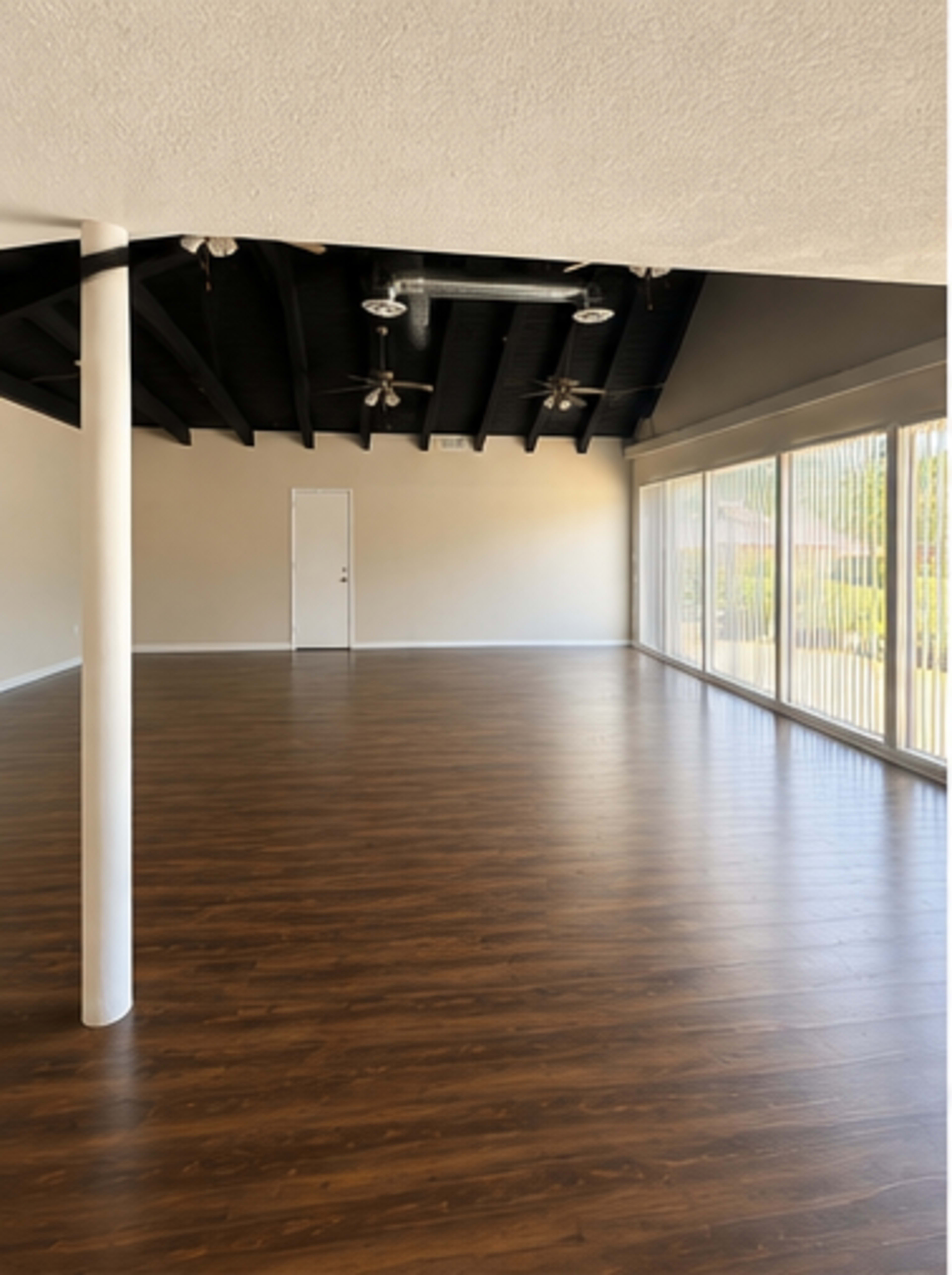 The image shows a spacious, empty room with wooden flooring, high black-beamed ceilings, and large windows that let in natural light.
