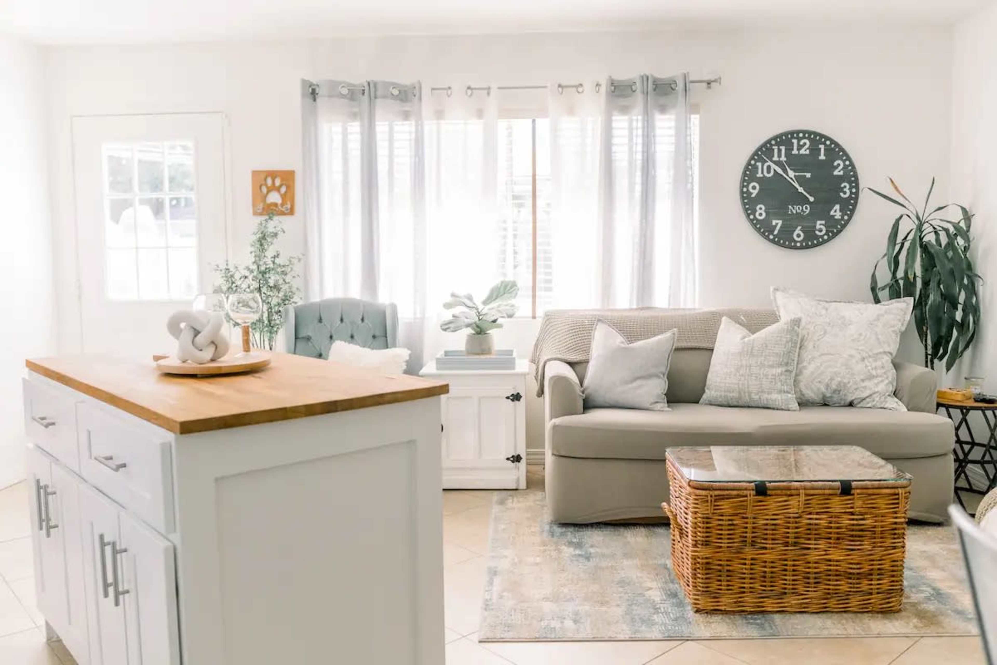 A bright living room features a beige sofa, a wooden coffee table, and a large wall clock, with natural light coming through sheer curtains.