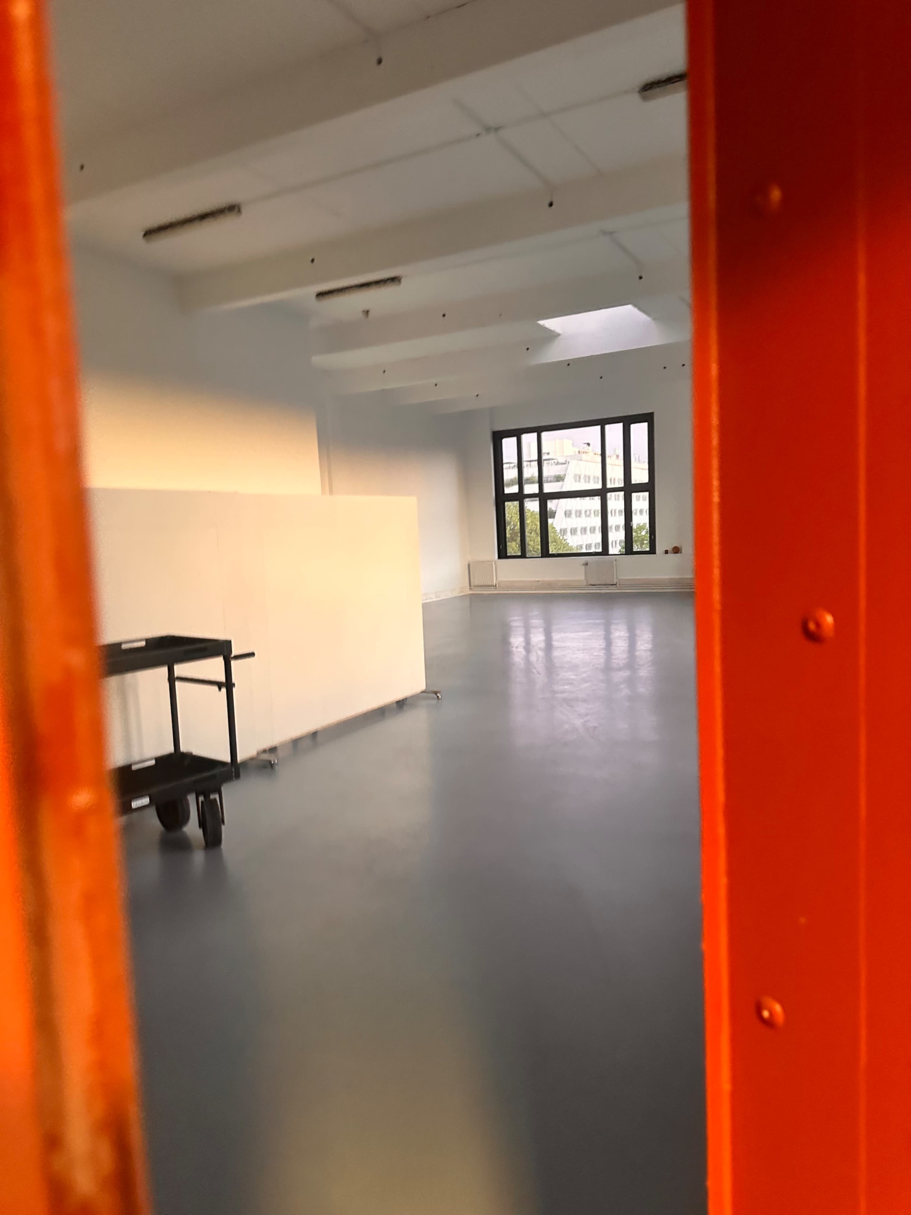 A brightly lit room with white walls and a large window, viewed through an open red door.