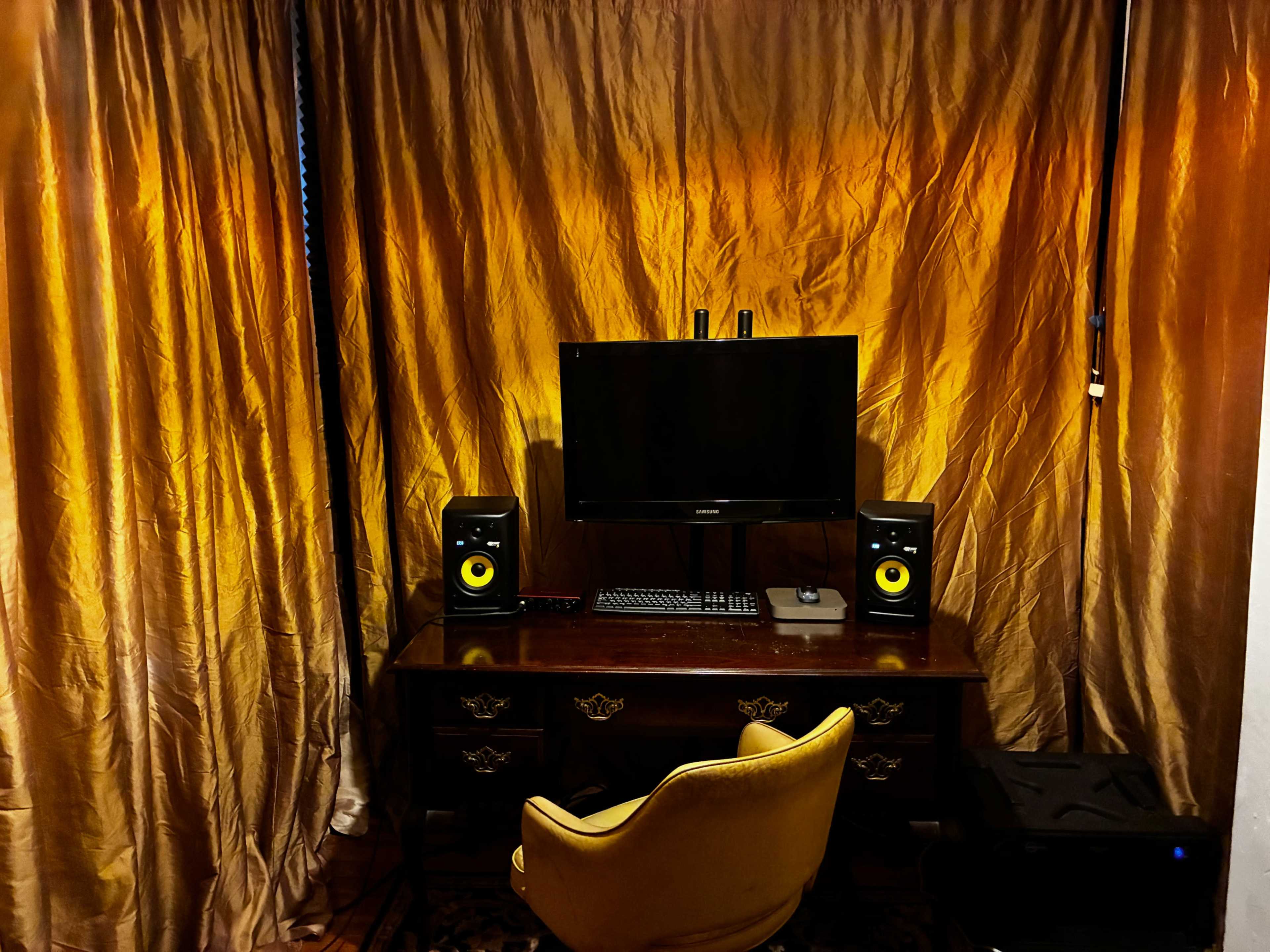 The image shows a workspace with a black computer monitor, two audio speakers, a keyboard, and an antique wooden desk, all set against a backdrop of golden curtains.