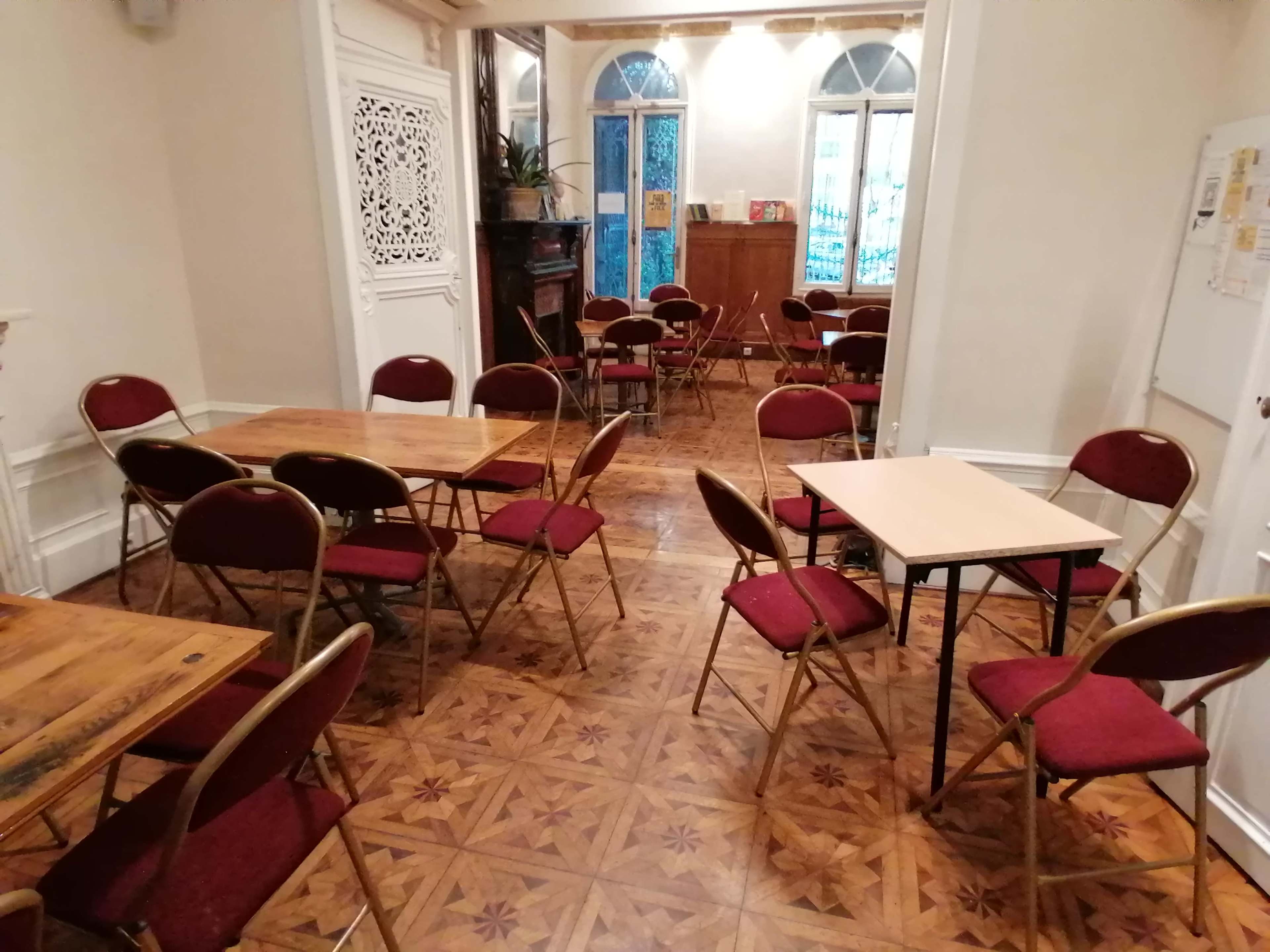 The image shows a room with several wooden and folding tables surrounded by chairs, featuring ornate wooden flooring and large windows.
