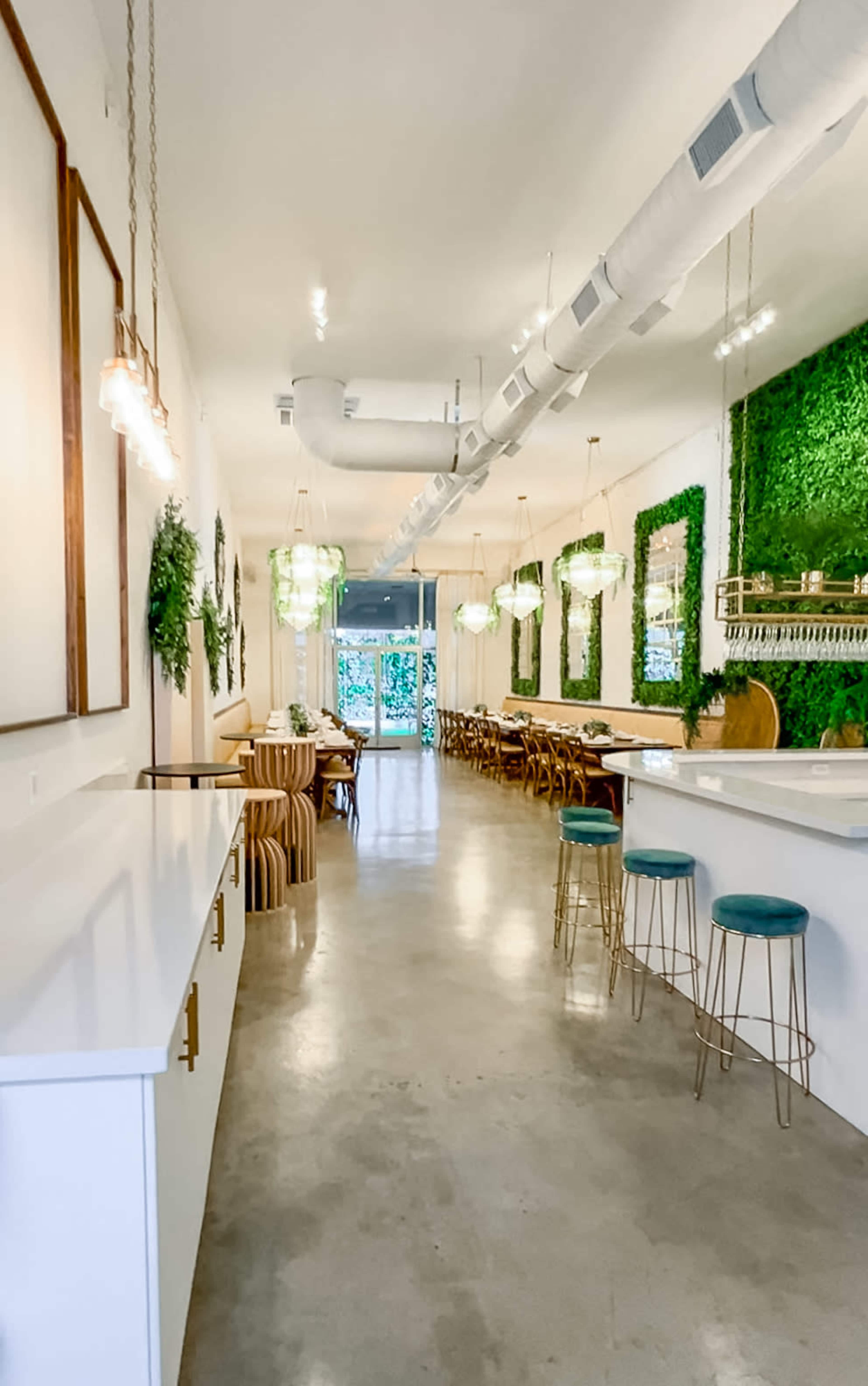 The image shows a modern restaurant interior with a concrete floor, green wall decor, and a mix of seating arrangements, including bar stools and wooden chairs.