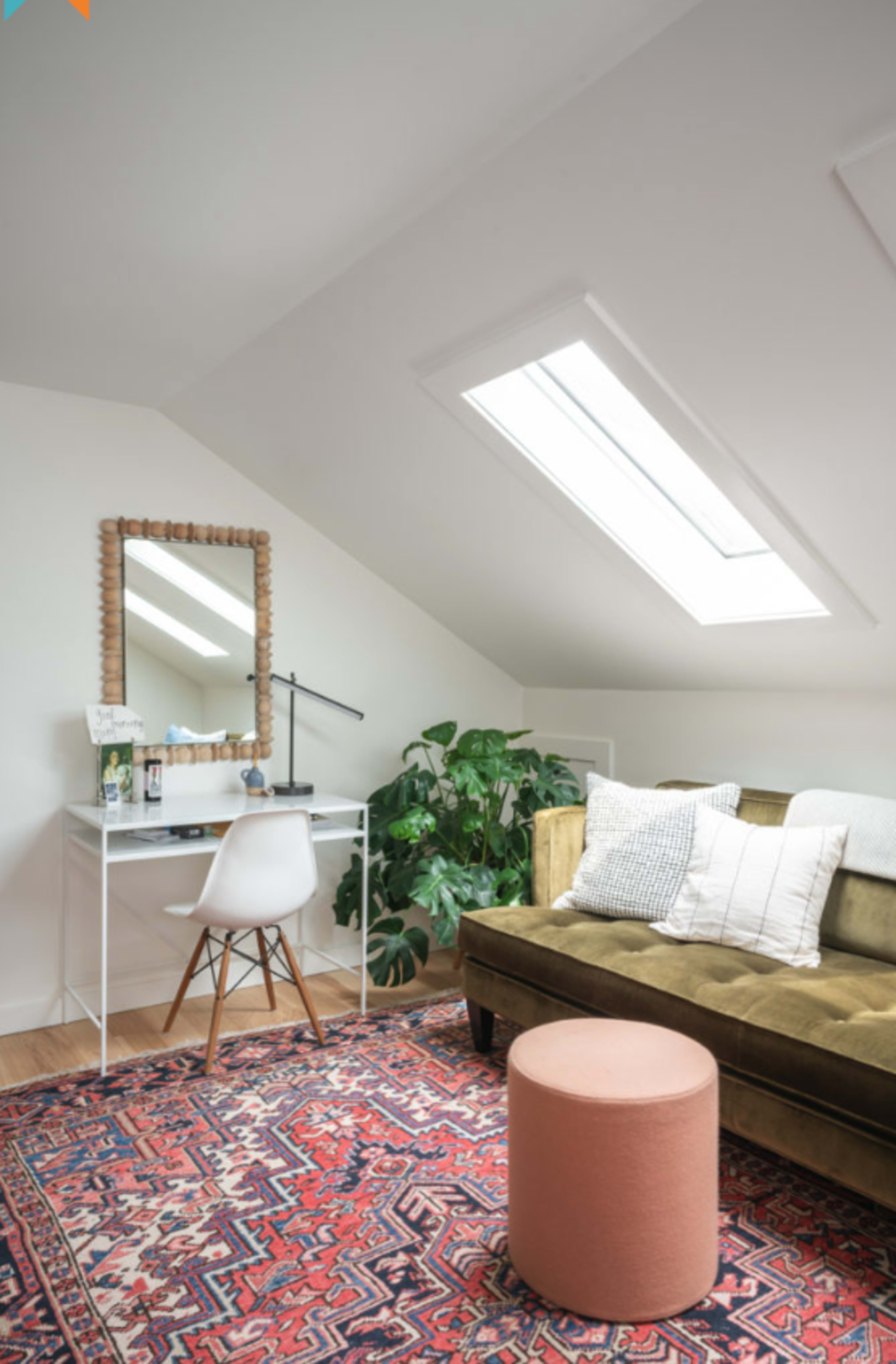 The image shows a cozy attic space featuring a sofa, a desk with a lamp, a round pink ottoman, a large mirror, and a potted plant under a sloped ceiling with skylights.