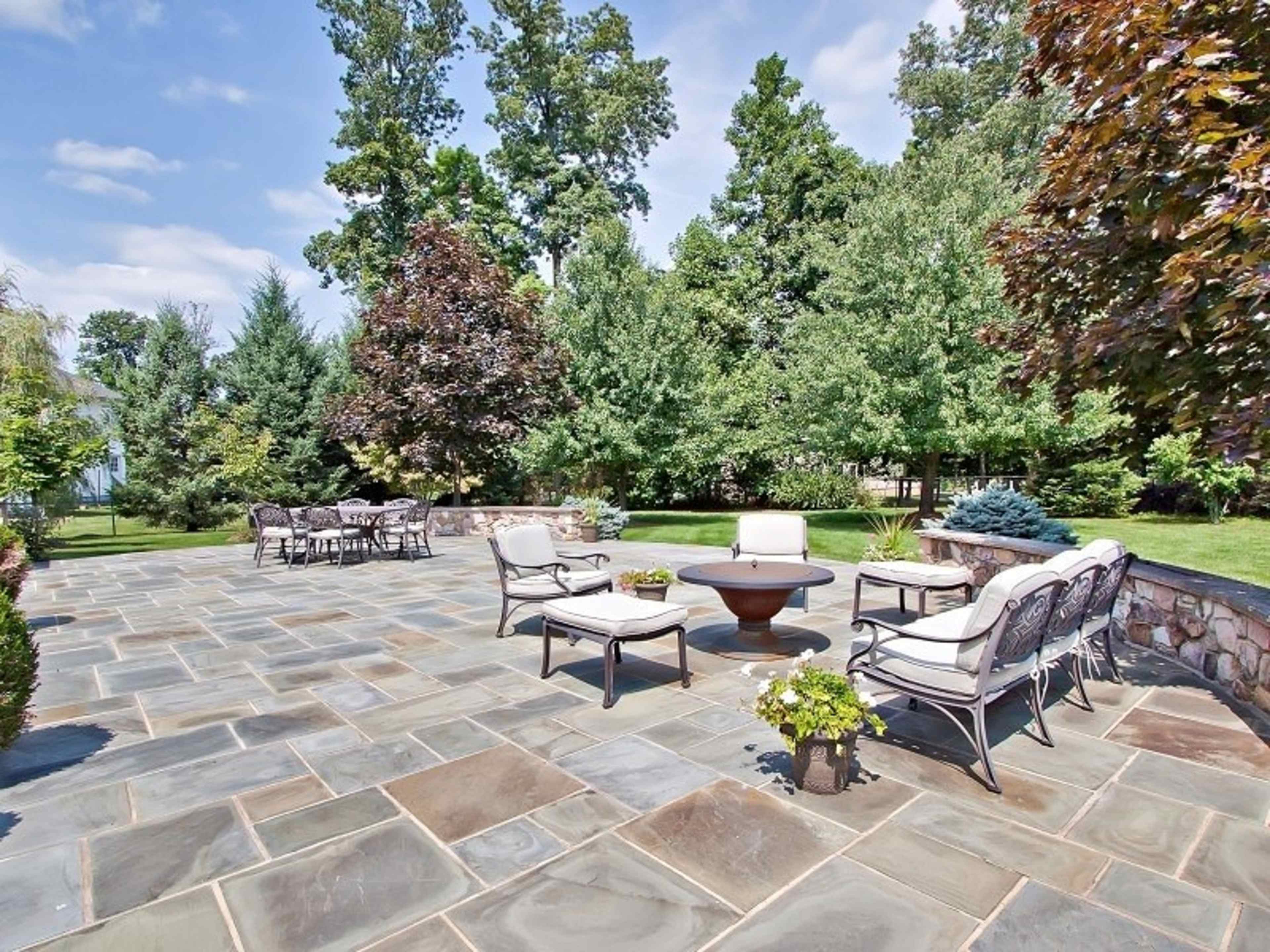 A stone patio features several seating arrangements, including chairs and a fire pit, surrounded by trees and greenery.