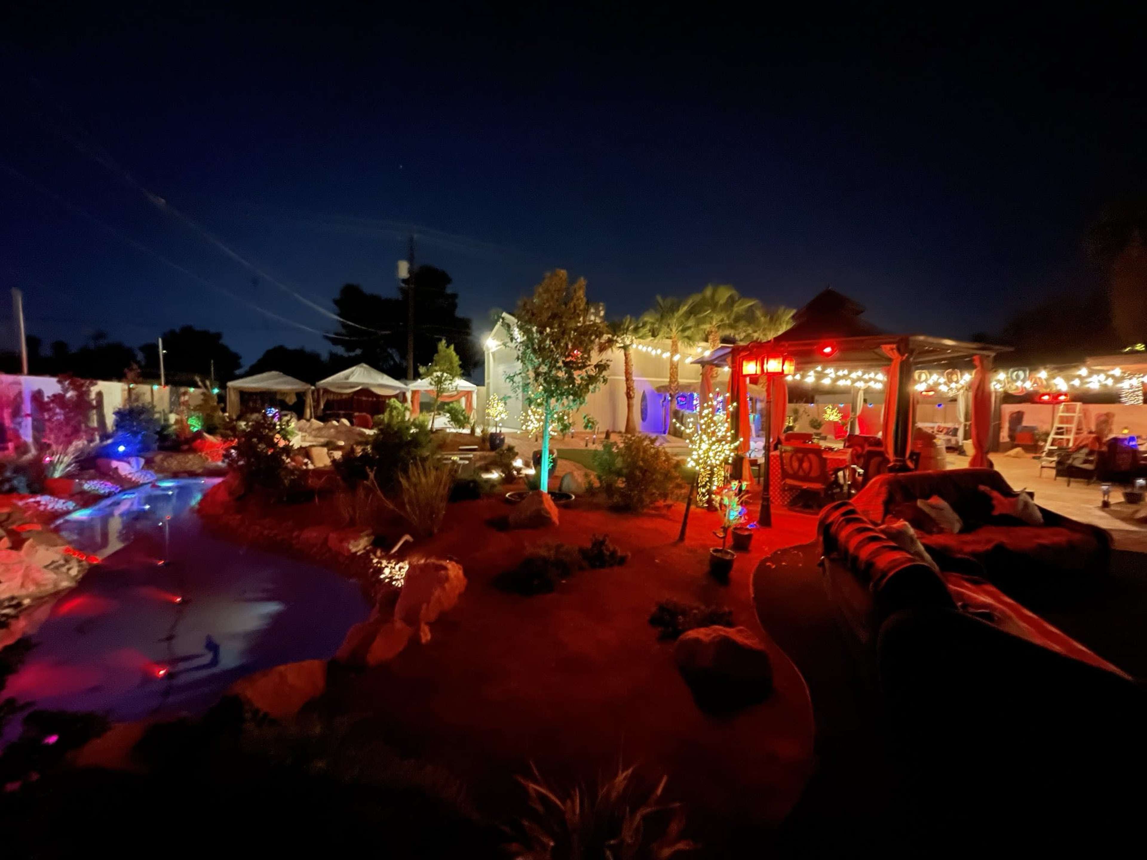 A backyard scene is illuminated with colorful lights at night, featuring a small water feature, seating areas, and decorative plants.