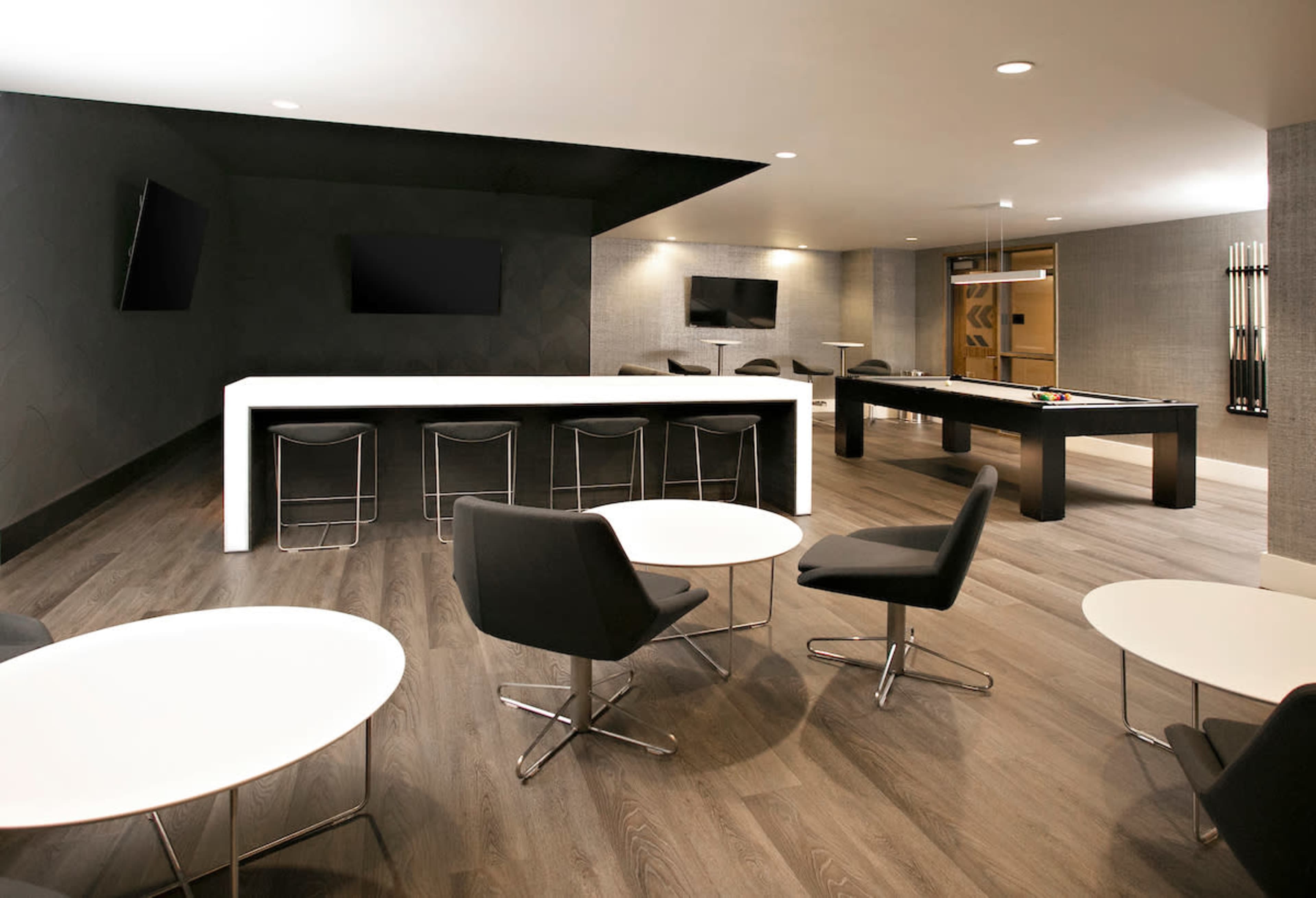 The image shows a modern lounge area featuring a bar, several round tables, and a billiards table against a sleek, dark wall.