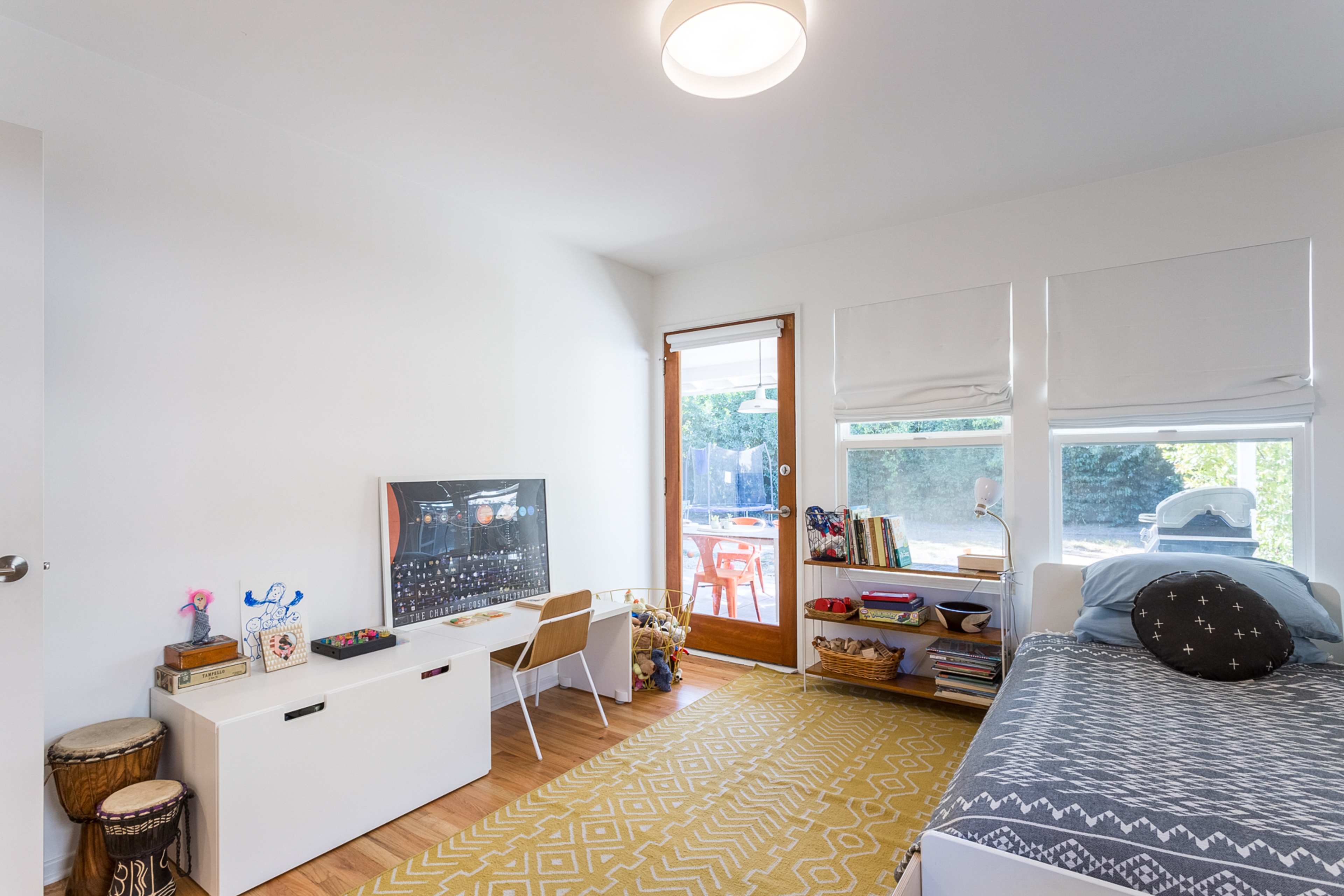 A children's bedroom features a bed, a white storage unit, and large windows leading to a backyard.