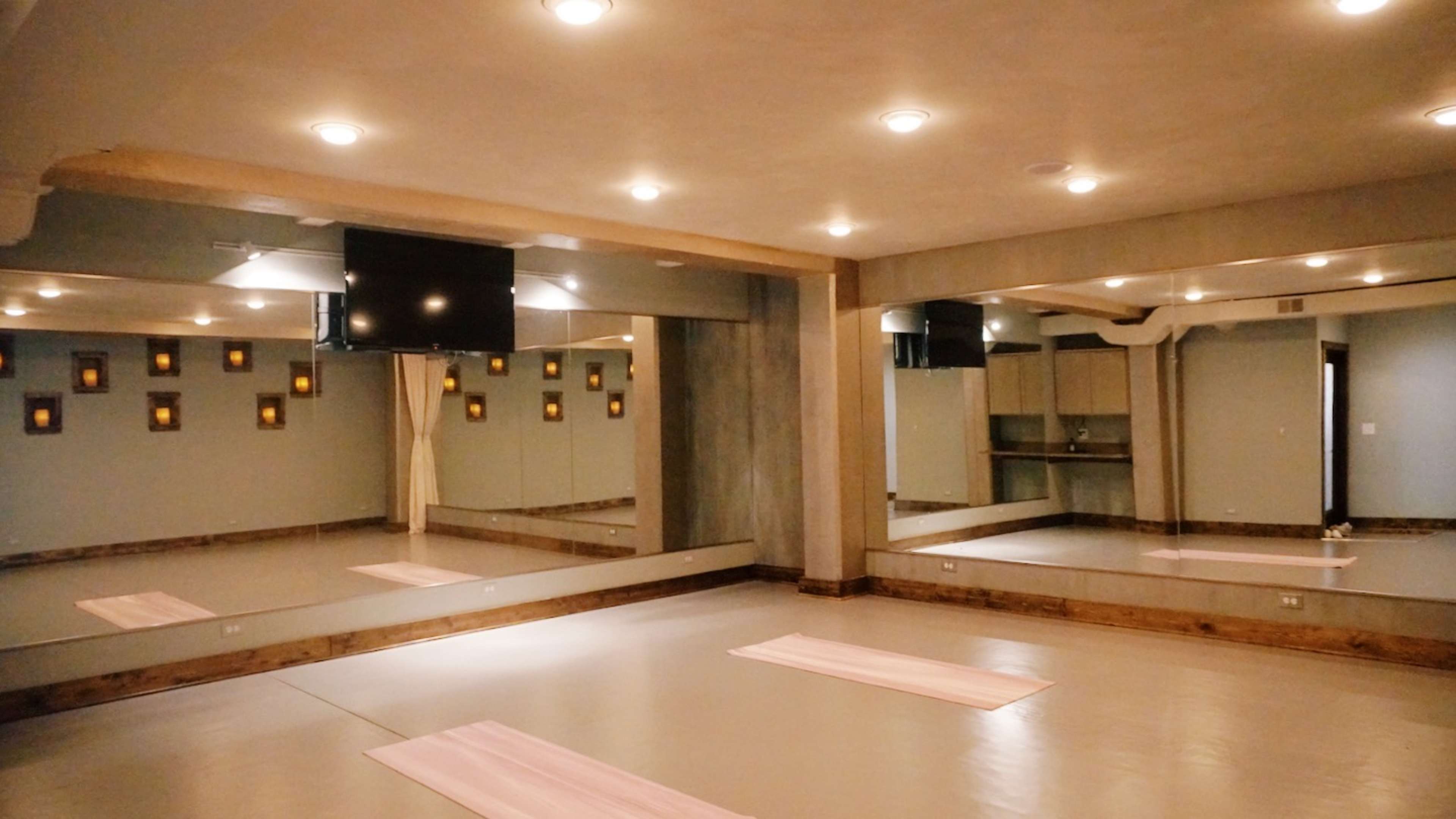 A spacious yoga studio with mirrors on one wall, soft lighting, and two pink mats laid out on the floor.