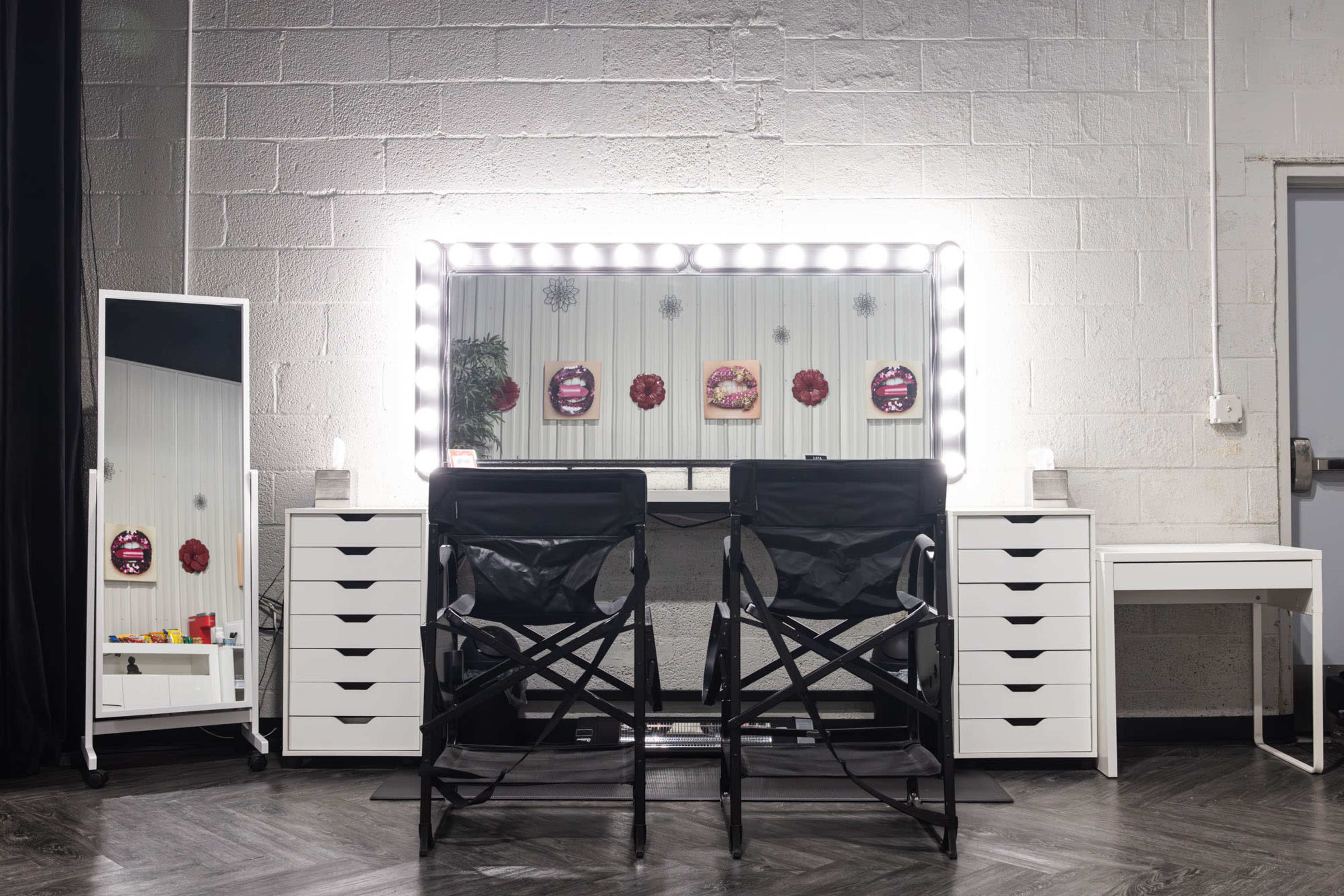 A brightly lit makeup station features two black director's chairs in front of a large mirror, surrounded by white storage cabinets and a standing mirror.