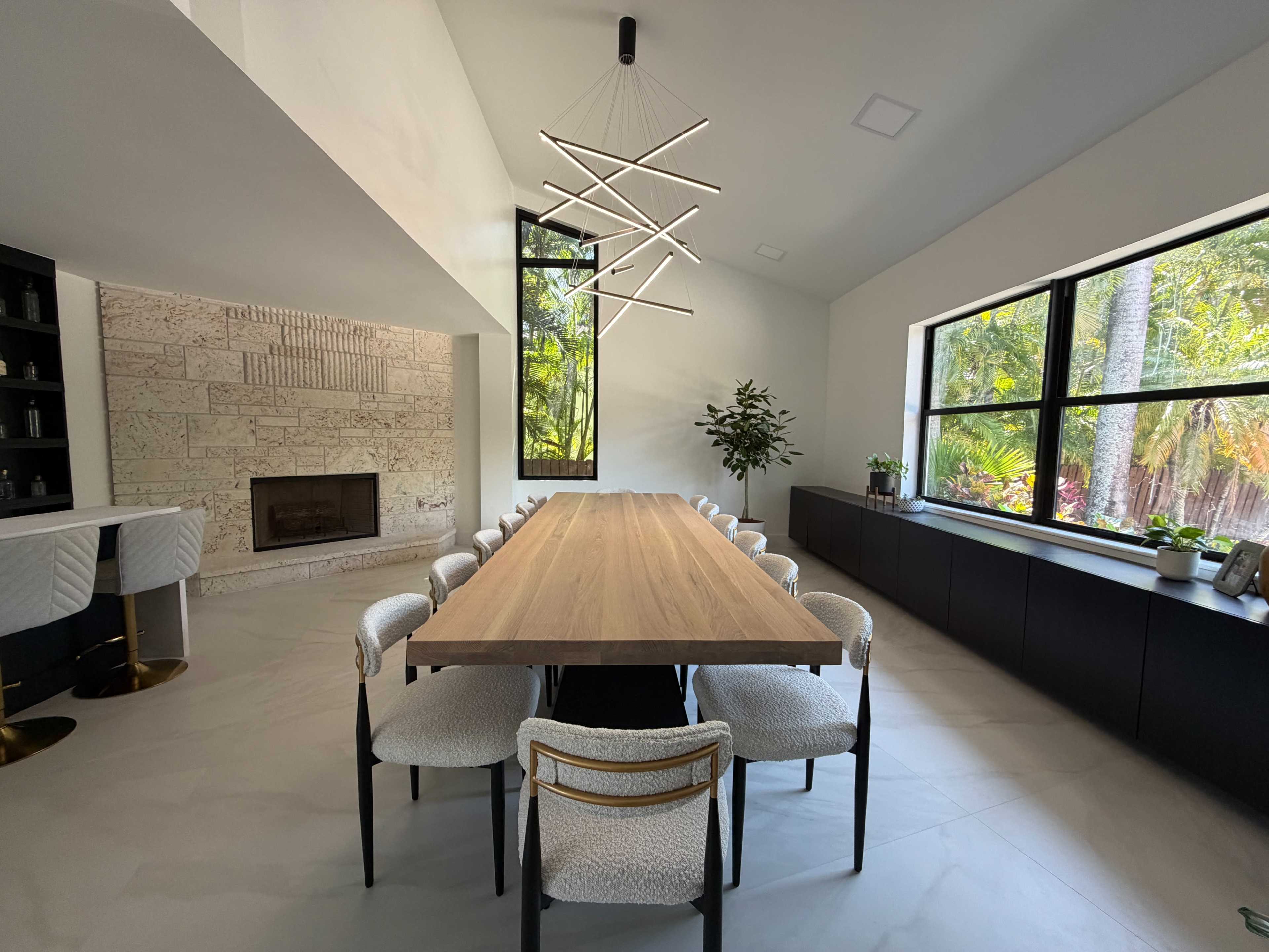 The image shows a modern dining room with a long wooden table surrounded by upholstered chairs, a minimalist light fixture overhead, and large windows allowing natural light to illuminate the space.