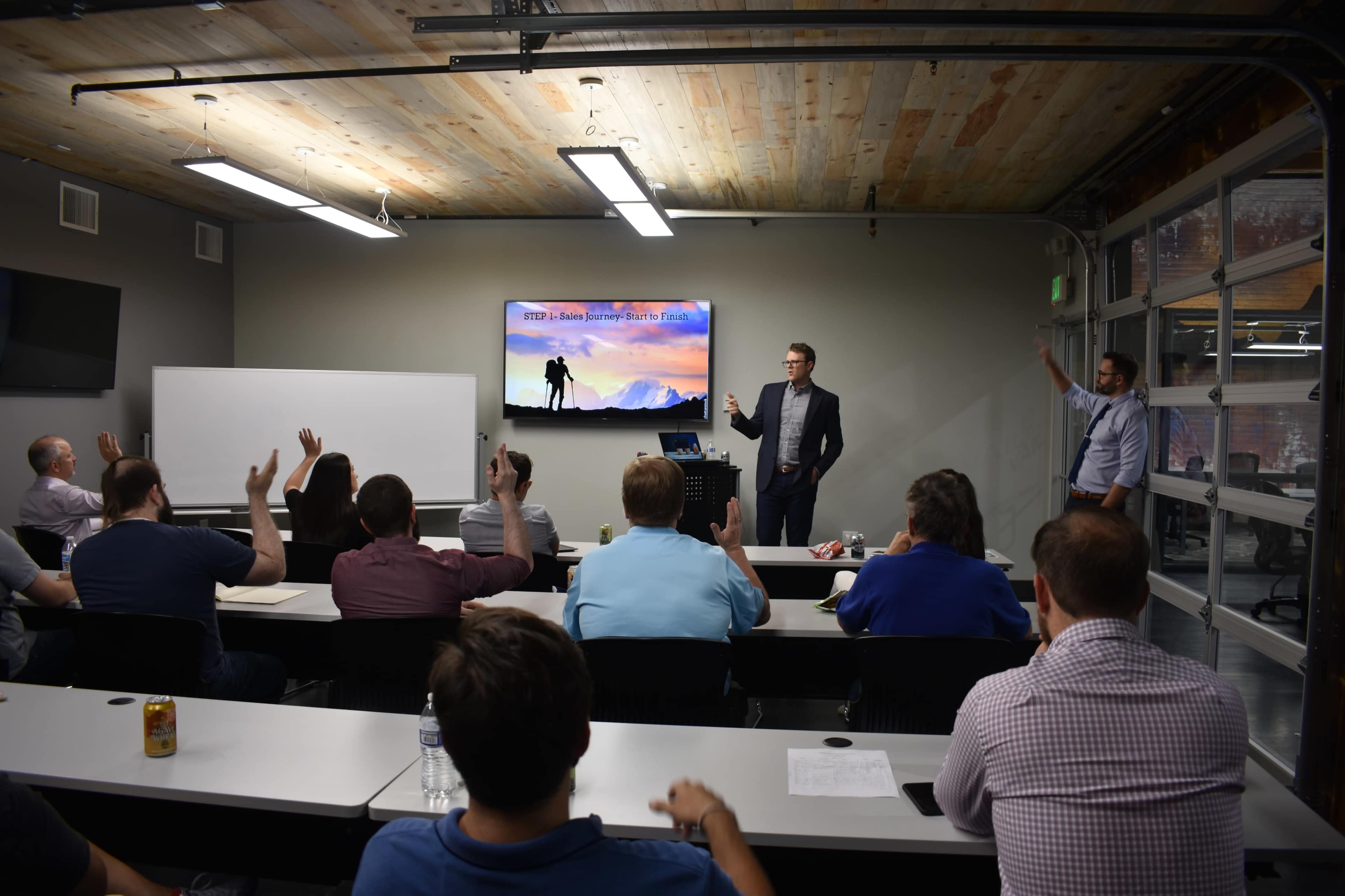 A speaker stands in front of an audience with raised hands in a modern classroom setting, showcasing a presentation on a screen.