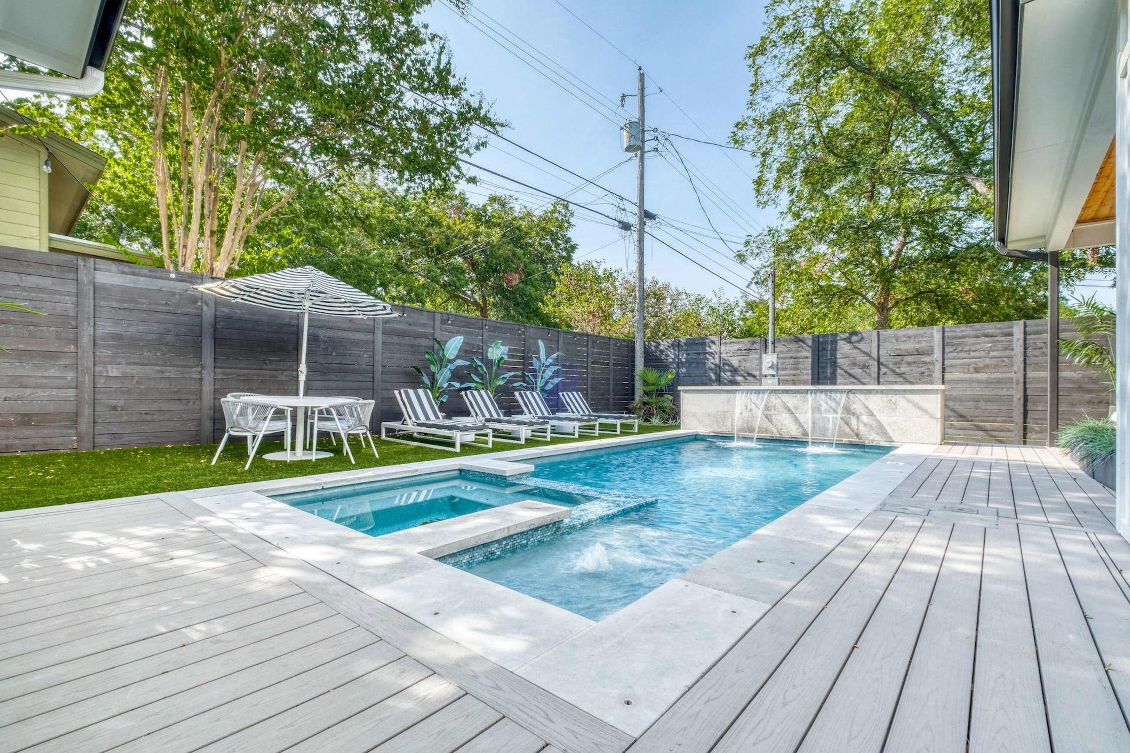 The image shows a backyard with a swimming pool, surrounded by lounge chairs and a small table under an umbrella, with a wooden deck and landscaped greenery.