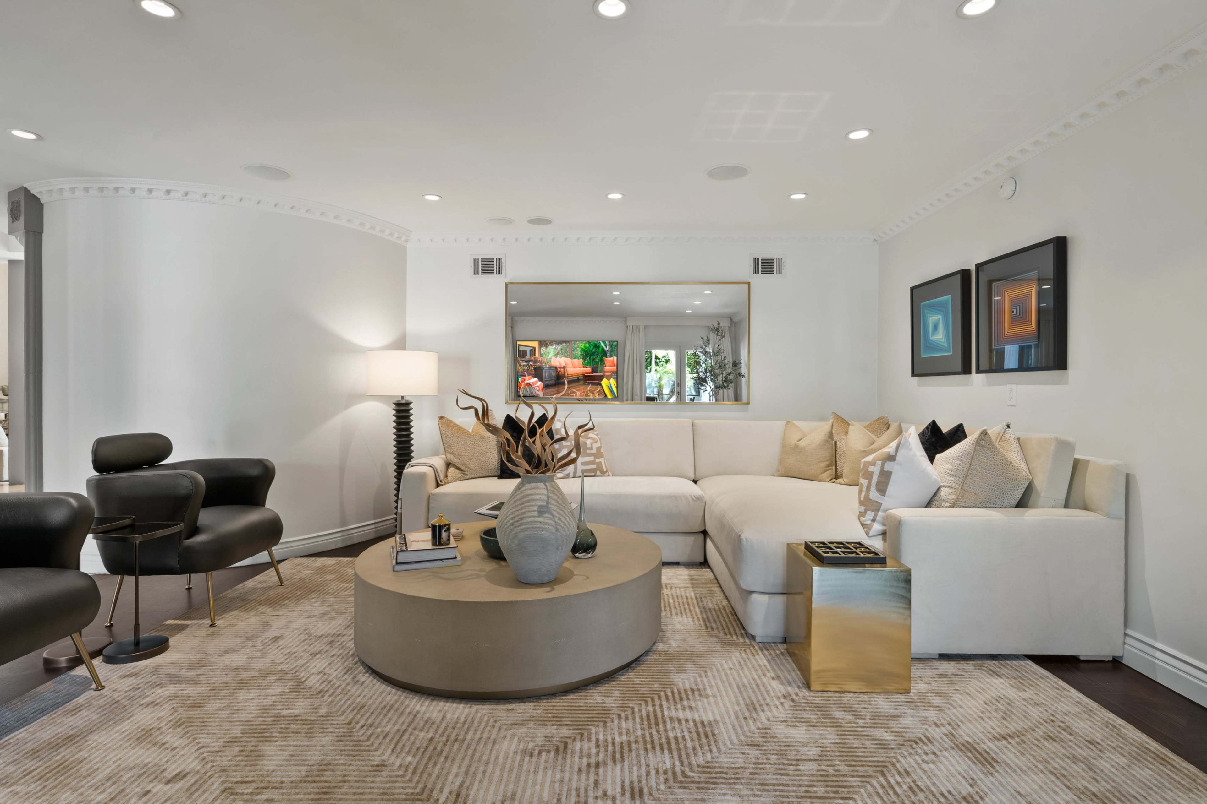 The image shows a modern living room featuring a white sectional sofa, a round coffee table, and two black chairs, with a mirror and artwork visible on the walls.