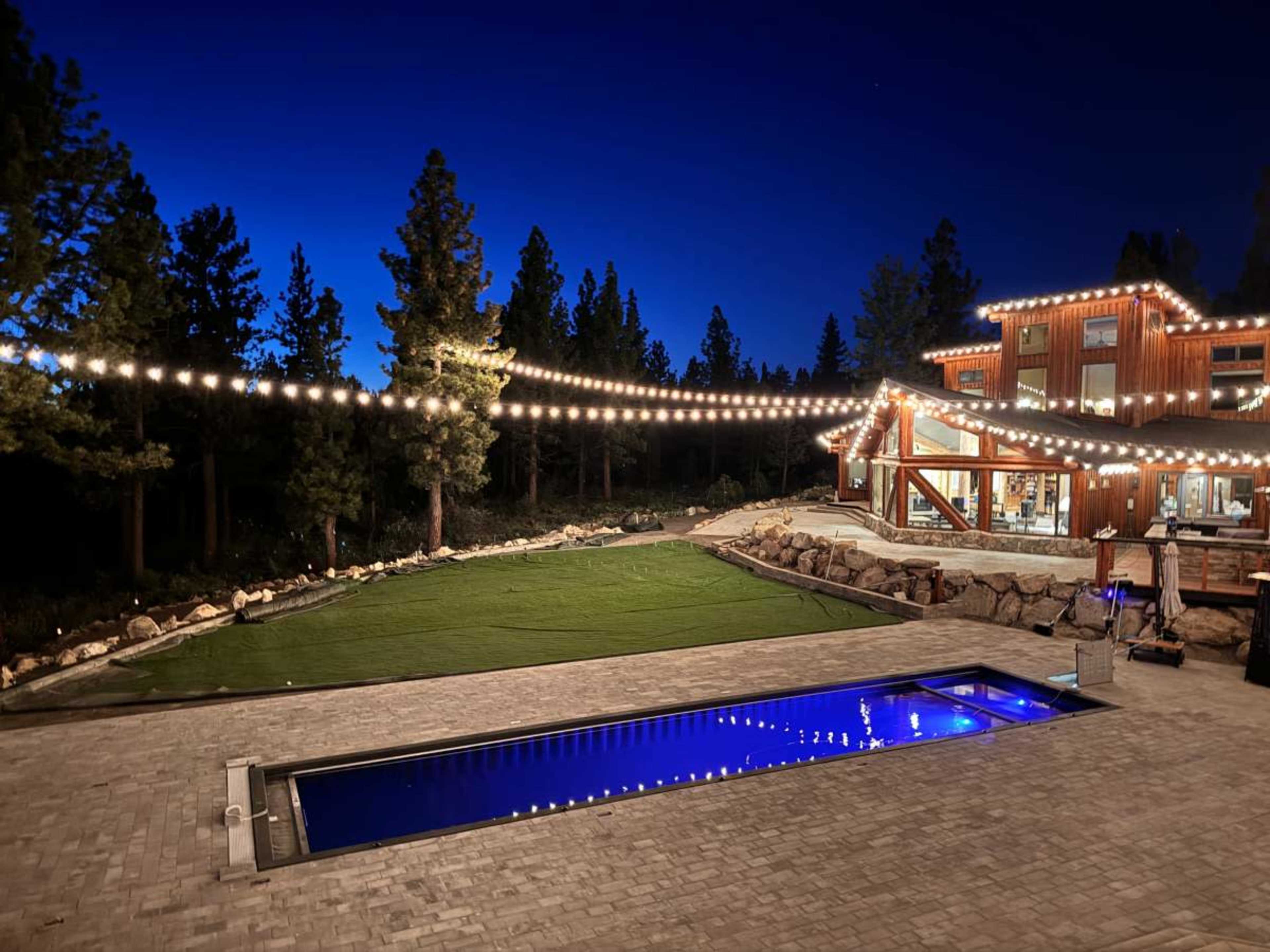 The image shows a backyard with a lit swimming pool, surrounded by trees and a modern wooden house, under a twilight sky.