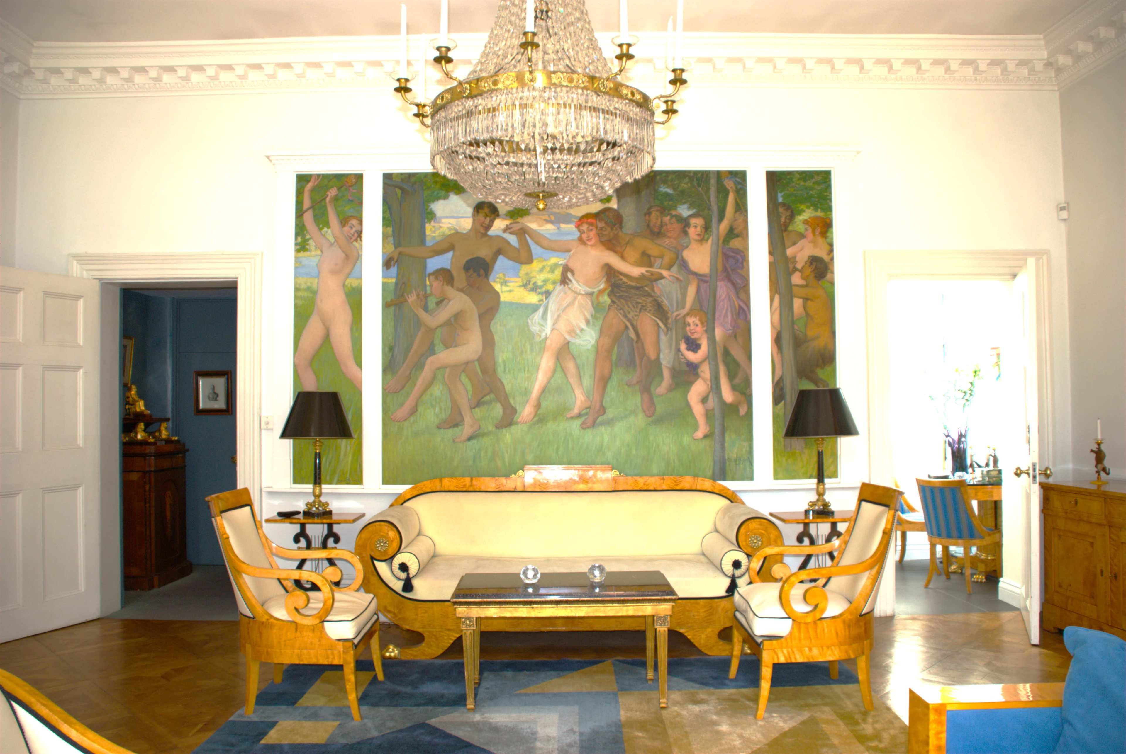 The image shows a spacious room furnished with a central curved sofa, two black lamps, and large murals depicting figures in a garden, illuminated by natural light from doorways on either side.